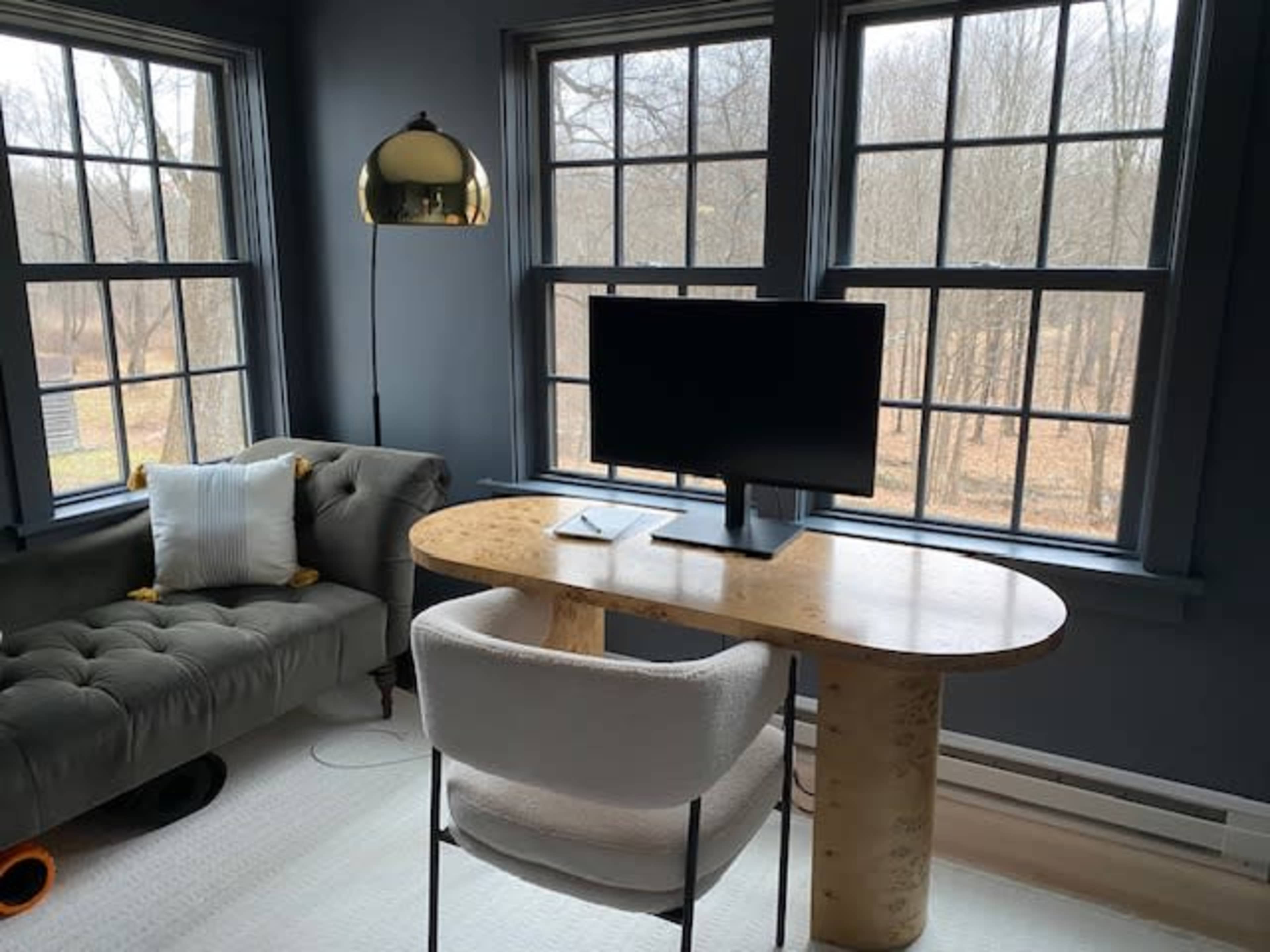 A modern workspace features a wooden desk with a computer monitor, set against large windows overlooking a wooded area, alongside a tufted sofa.