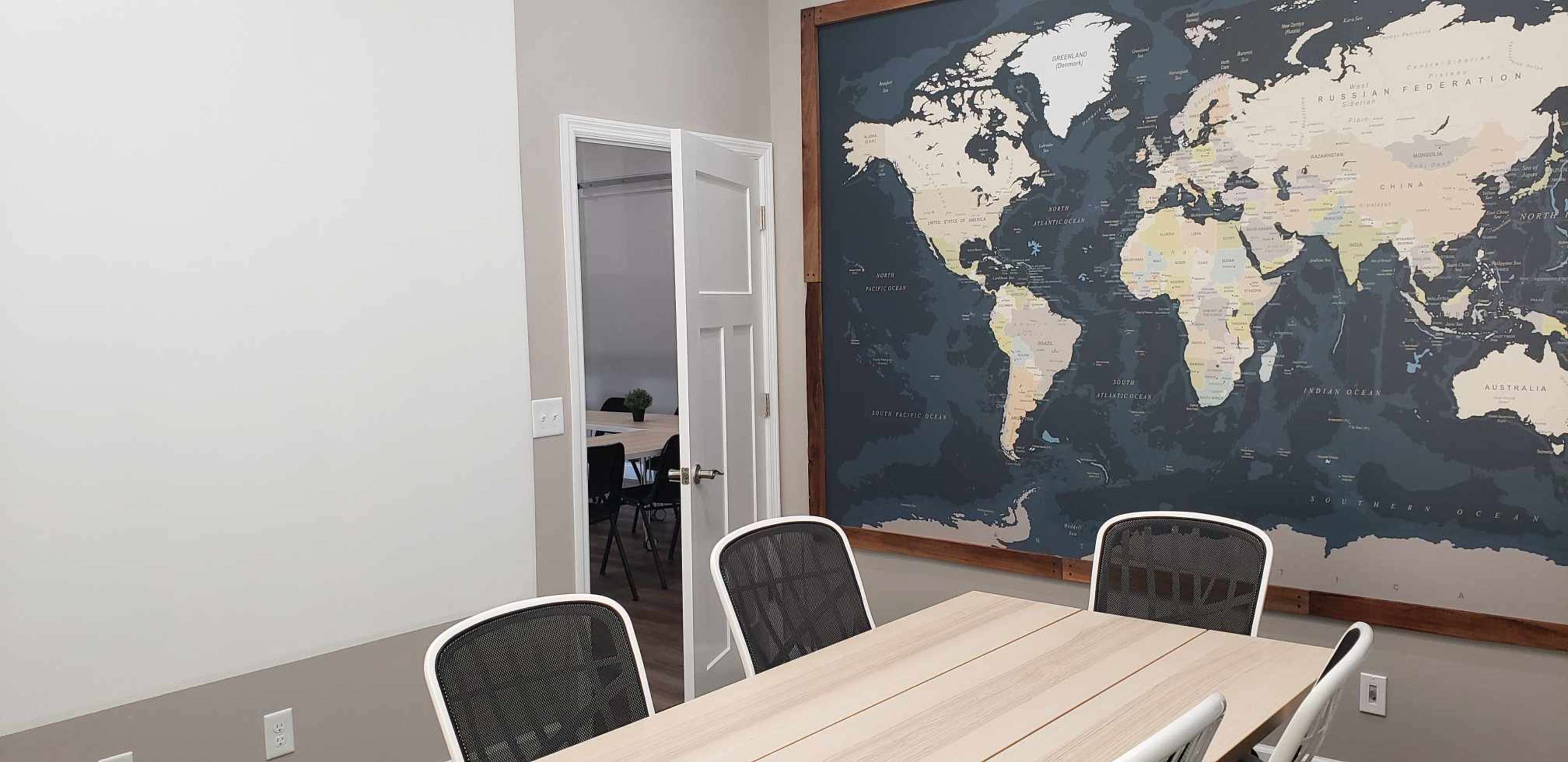 A meeting room features a large world map on the wall next to a wooden table and chairs, with an open door leading to another space.