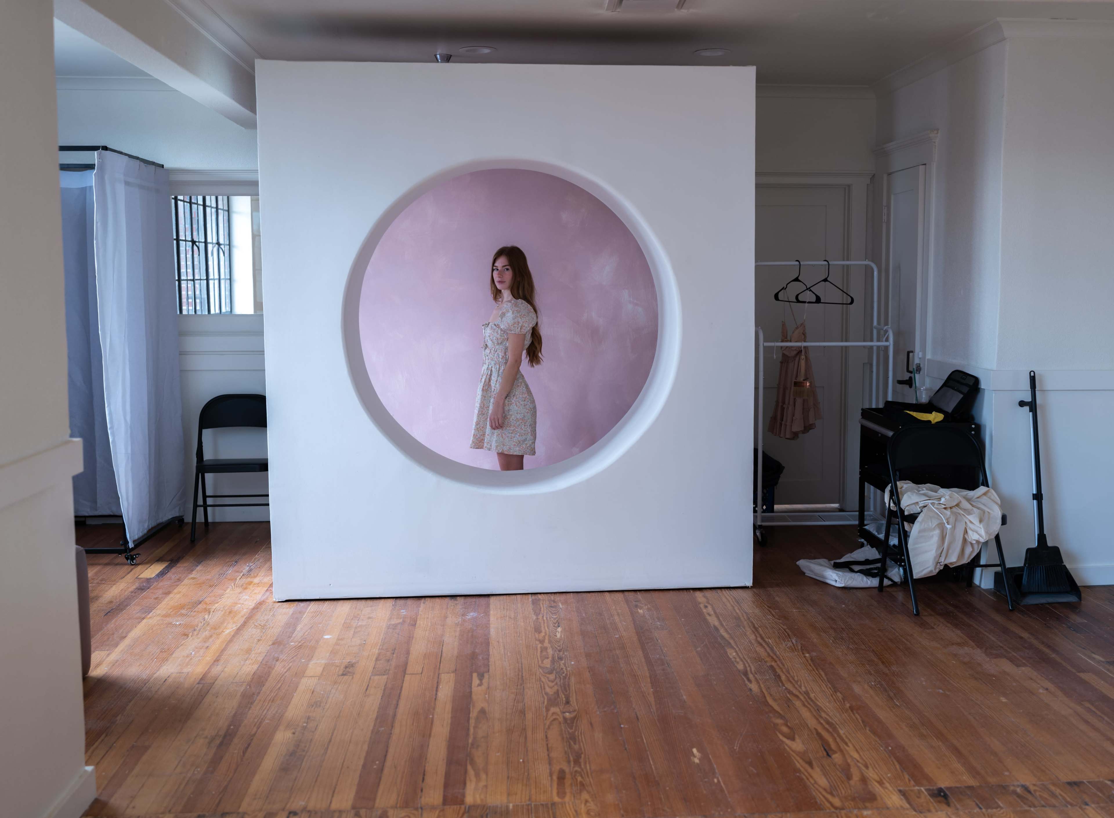 A young woman stands inside a circular cutout in a white wall, set against a pink backdrop in a room with wooden floors and minimal furnishings.