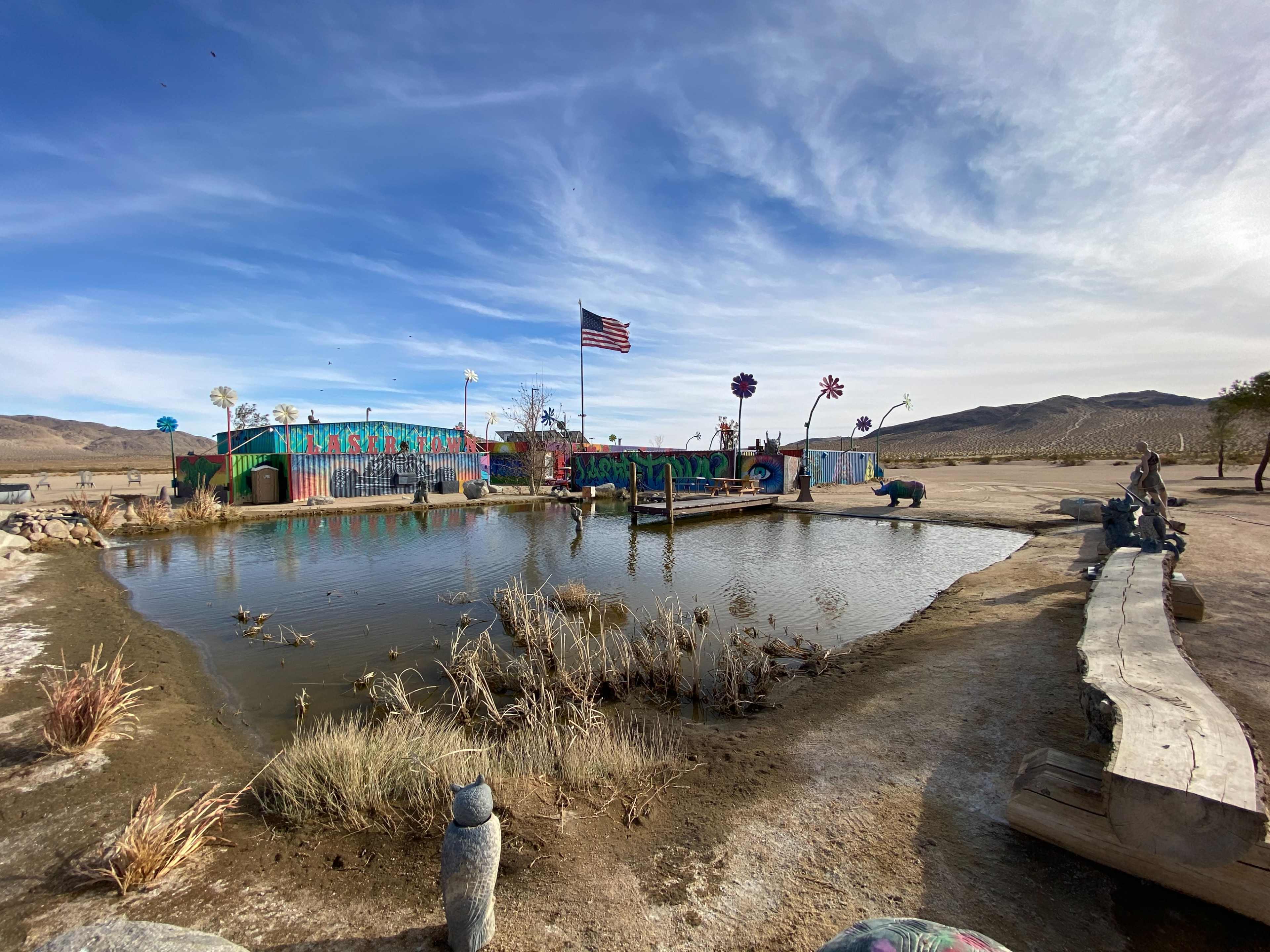 The image shows a colorful outdoor area with a pond, decorative structures, and an American flag in the background, set against a desert landscape.