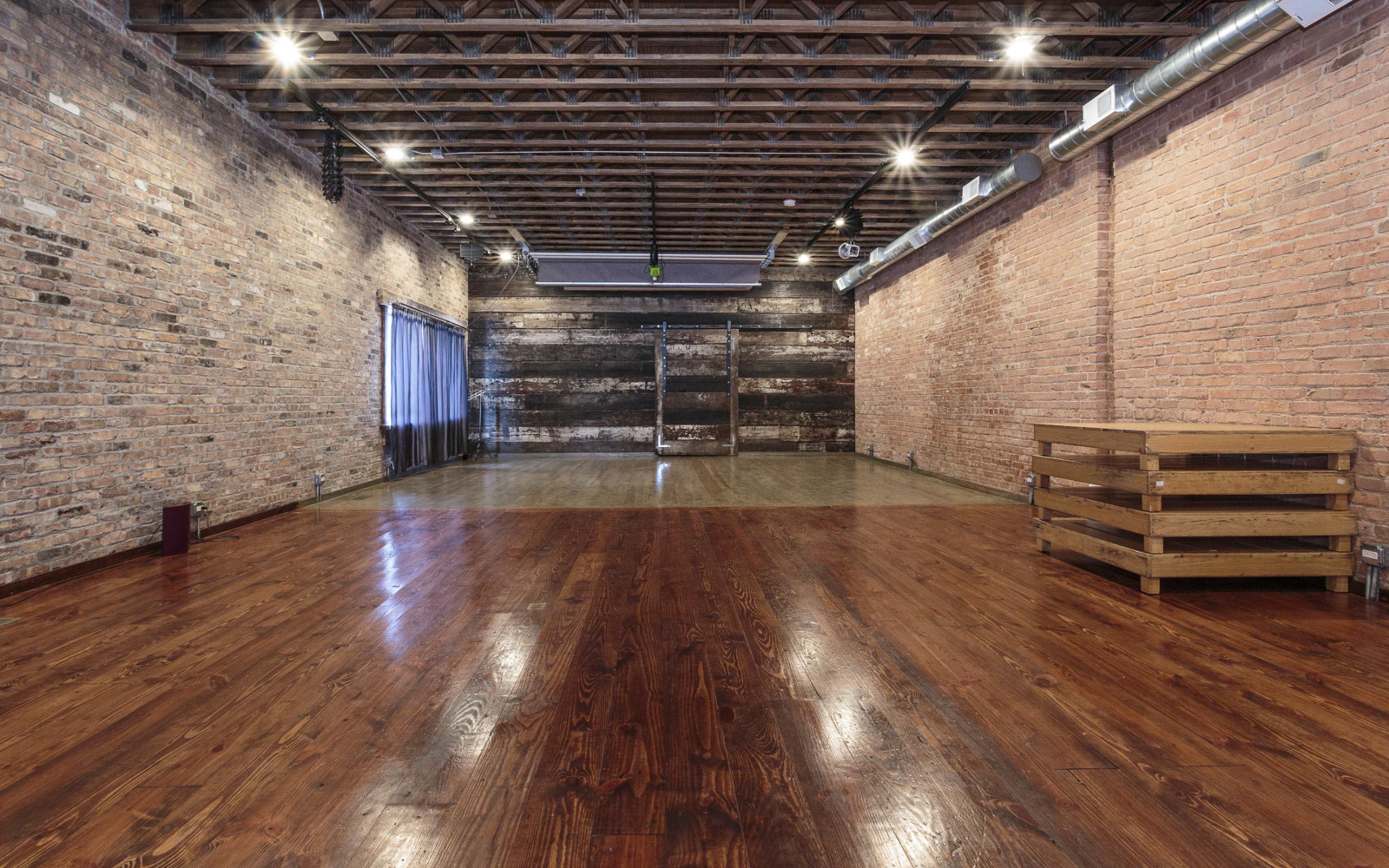 The image shows a spacious room with hardwood floors, exposed brick walls, and a wooden beam ceiling.