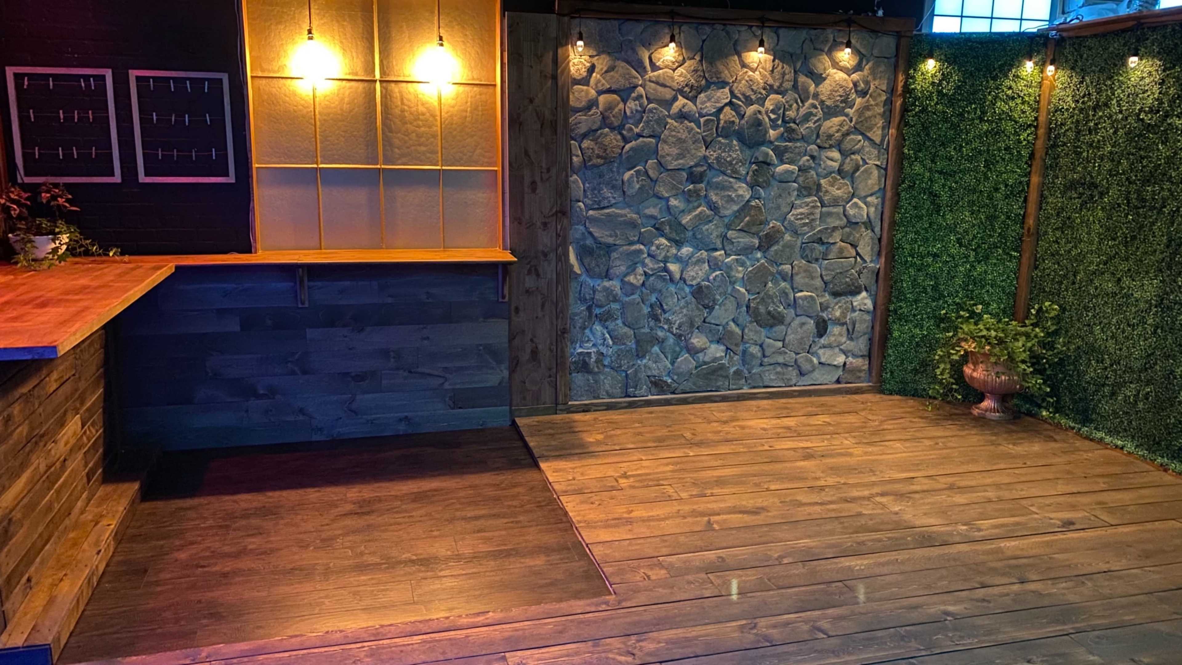 A cozy indoor space featuring a wooden floor, a stone wall, and warm lighting, with a bar area on the left and greenery on the right.