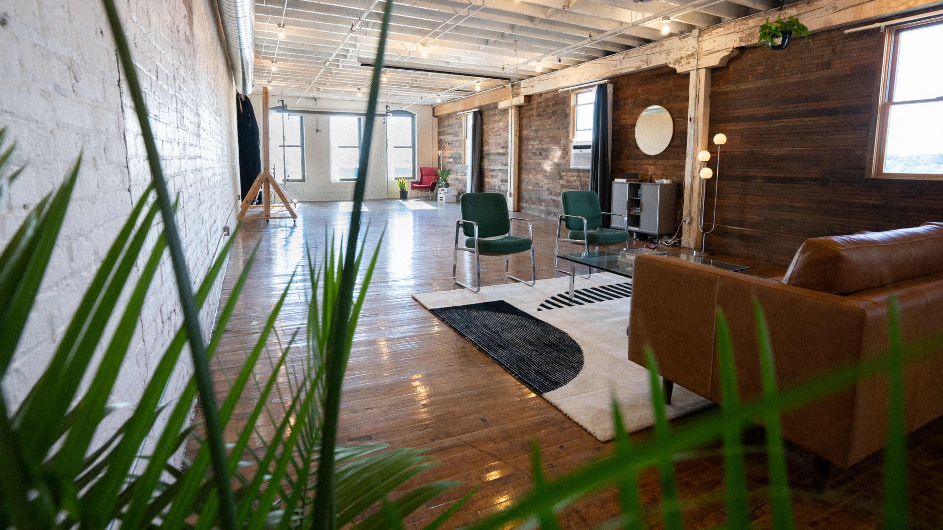 A spacious, open room with wooden flooring, featuring a brown sofa, two green chairs, a coffee table, and large windows that allow natural light to fill the space.