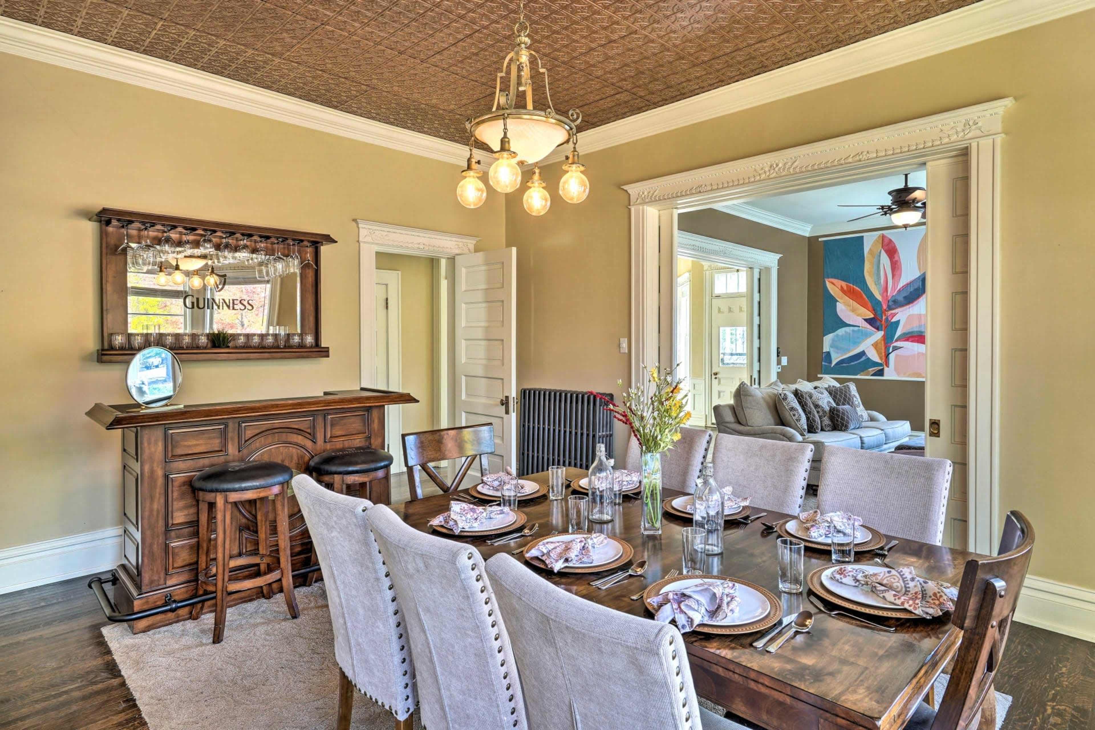 A dining area features a wooden table set for six with a bar in the corner, and a view of a living room through an adjacent doorway.