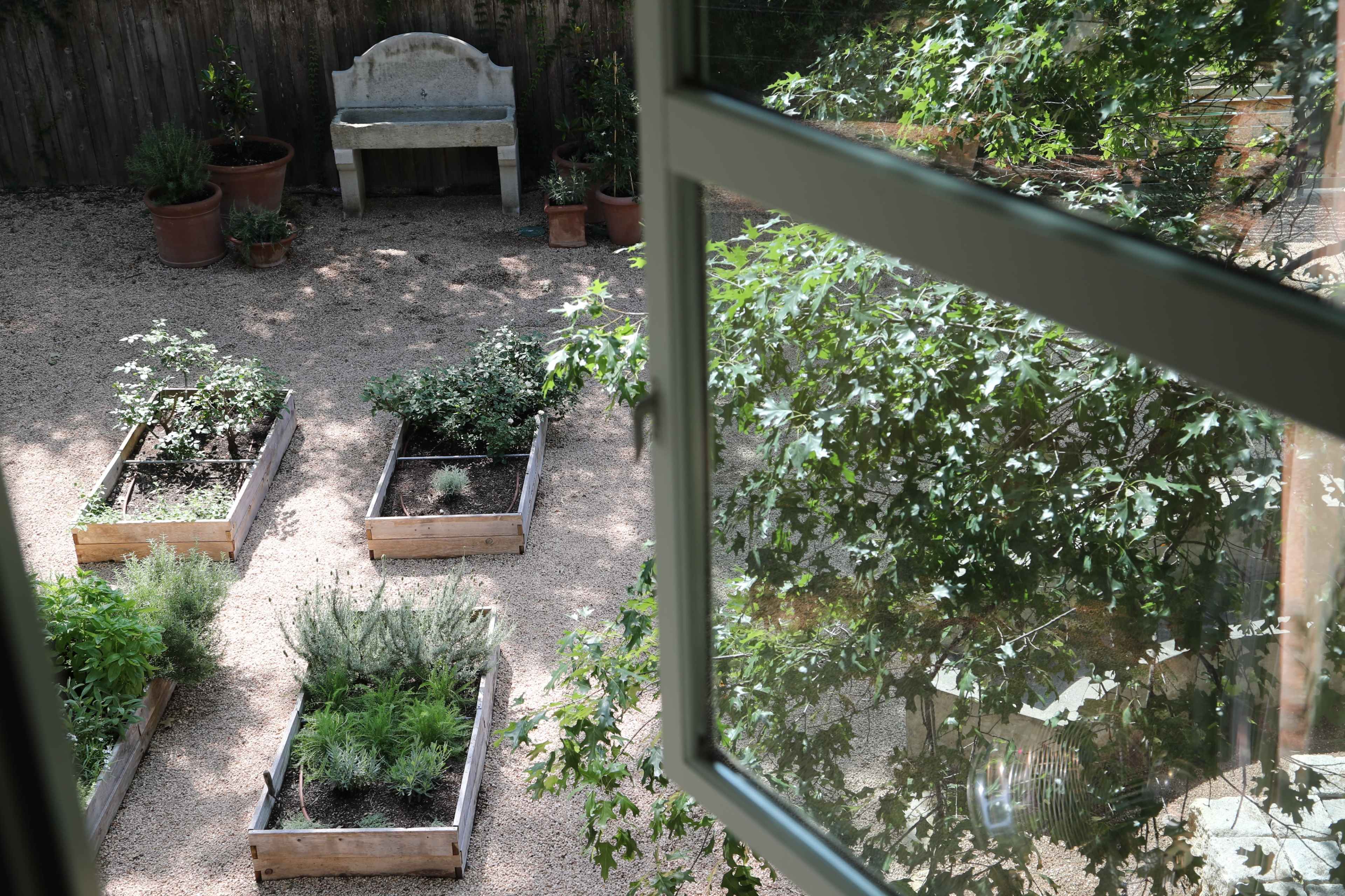 The image shows a view from a window overlooking a garden with raised wooden planters containing various plants and a stone bench in the background.
