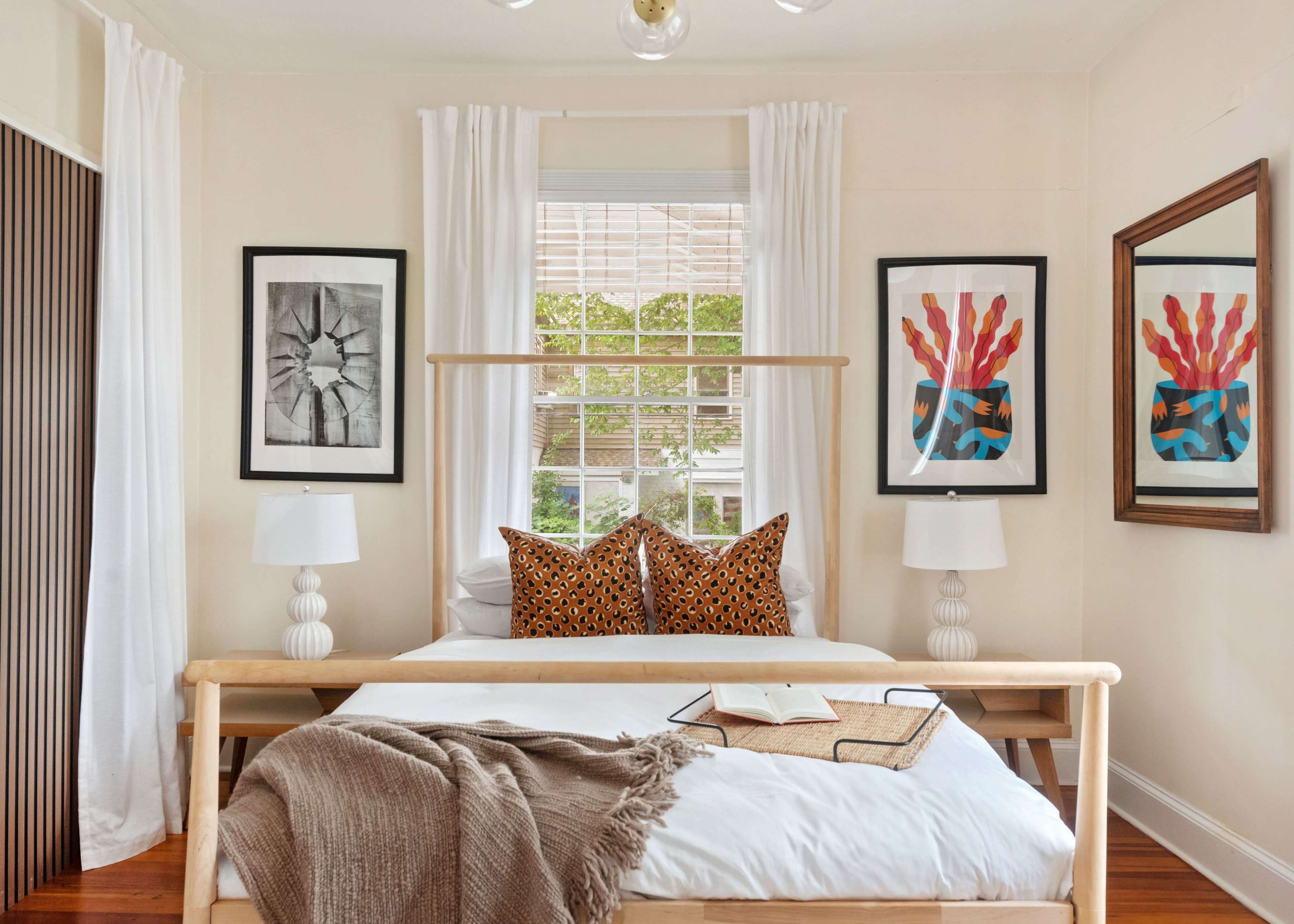 The image shows a cozy bedroom featuring a wooden bed with decorative pillows, two bedside lamps, framed artwork on the walls, and large windows with sheer curtains.