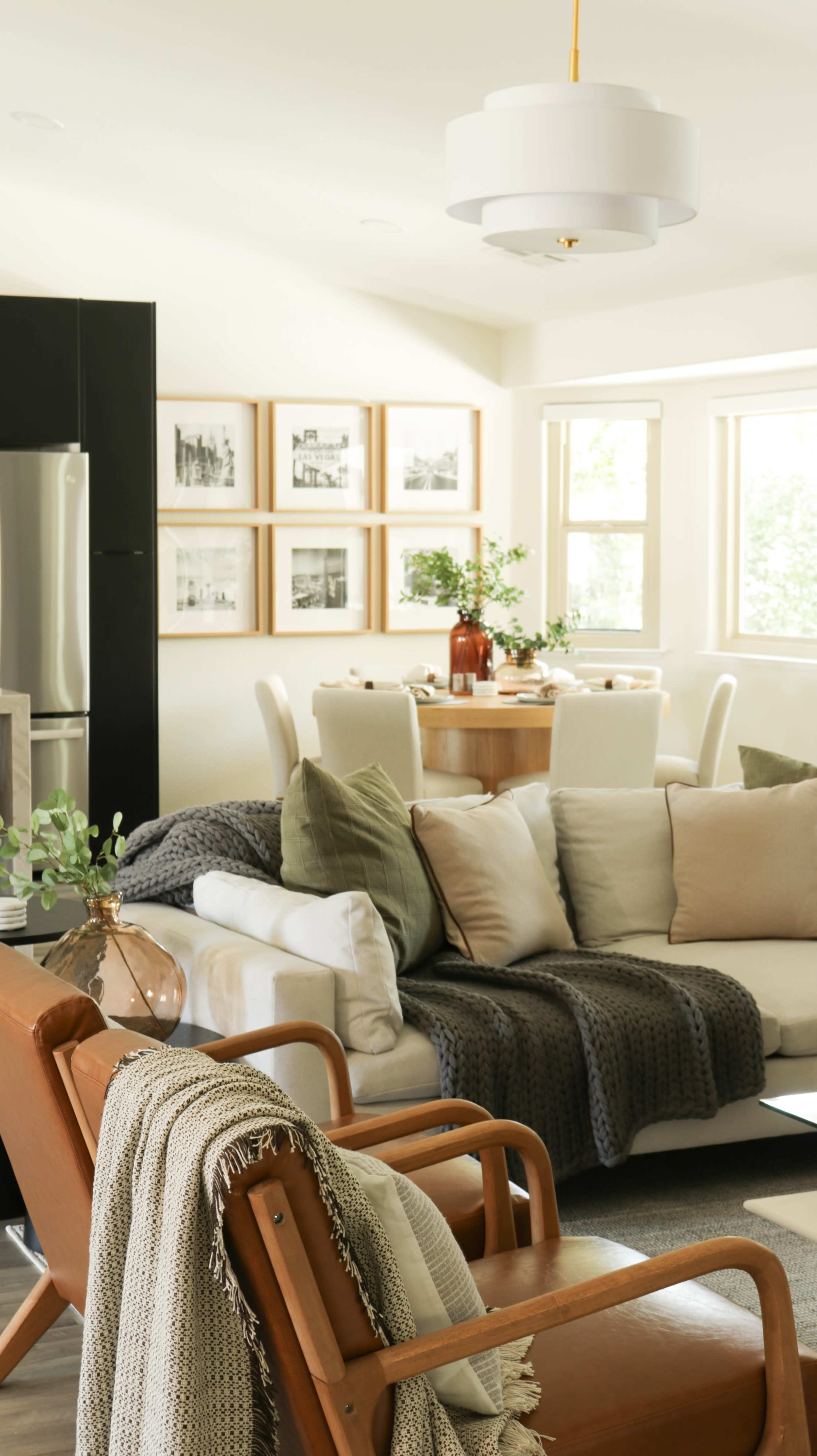 A modern living room features a beige sectional sofa with various sized cushions, a wooden coffee table, and a dining area with a round table and white chairs, all illuminated by natural light from large windows.