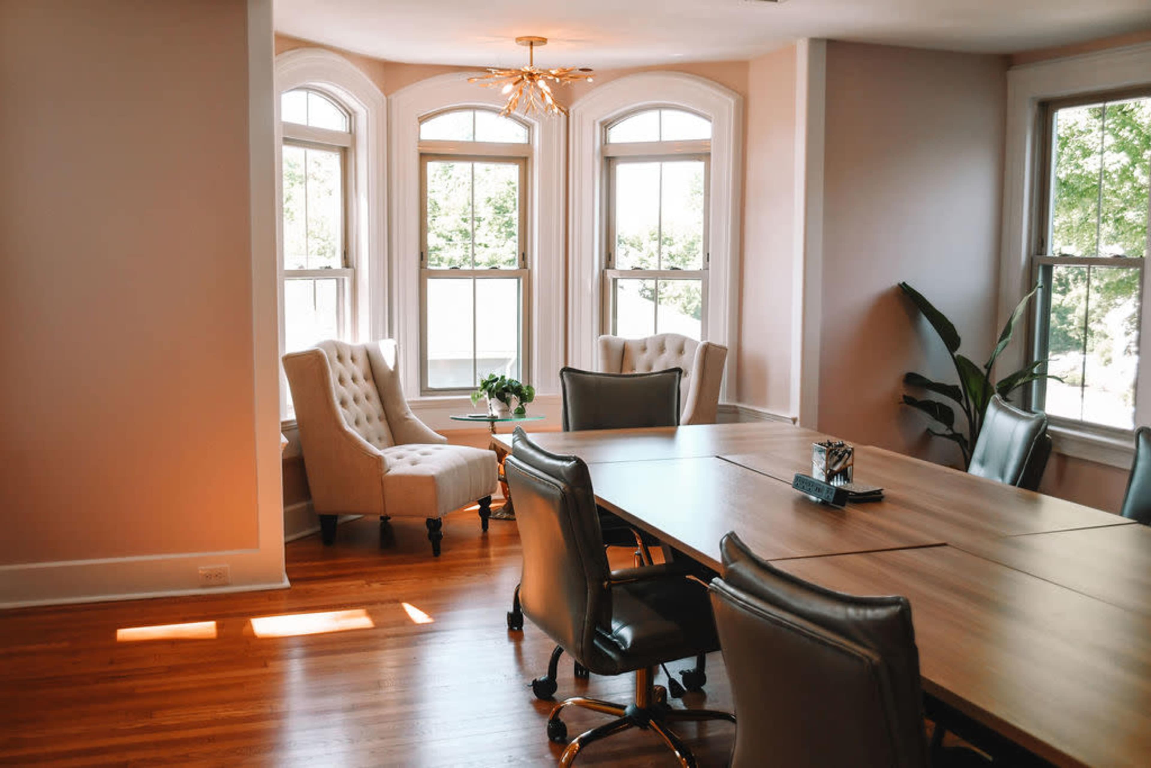 A modern conference room features a large wooden table surrounded by rolling chairs, with tall windows allowing natural light to fill the space.