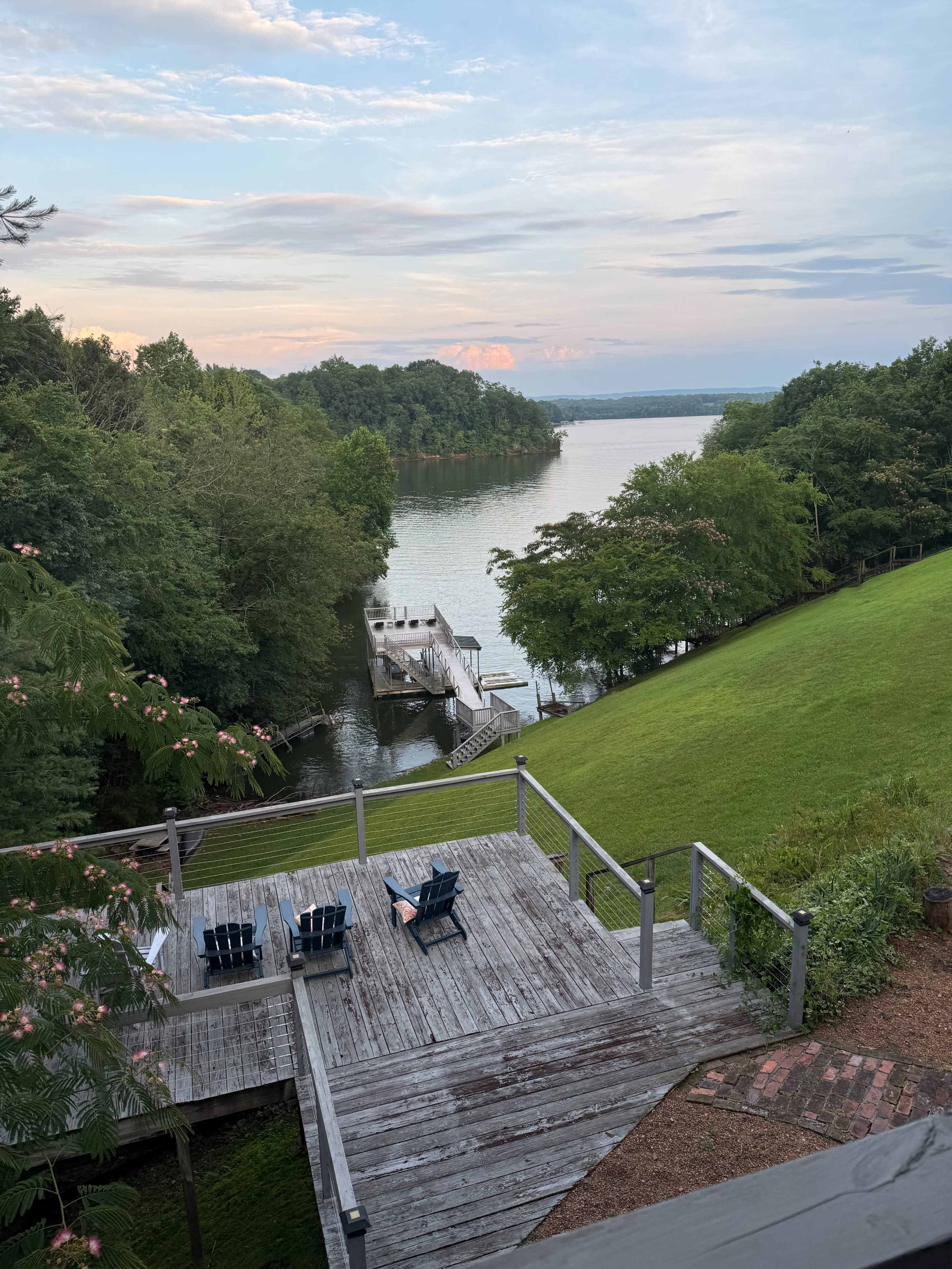 The image shows a wooden deck overlooking a calm river with a small dock and lush greenery surrounding the area.