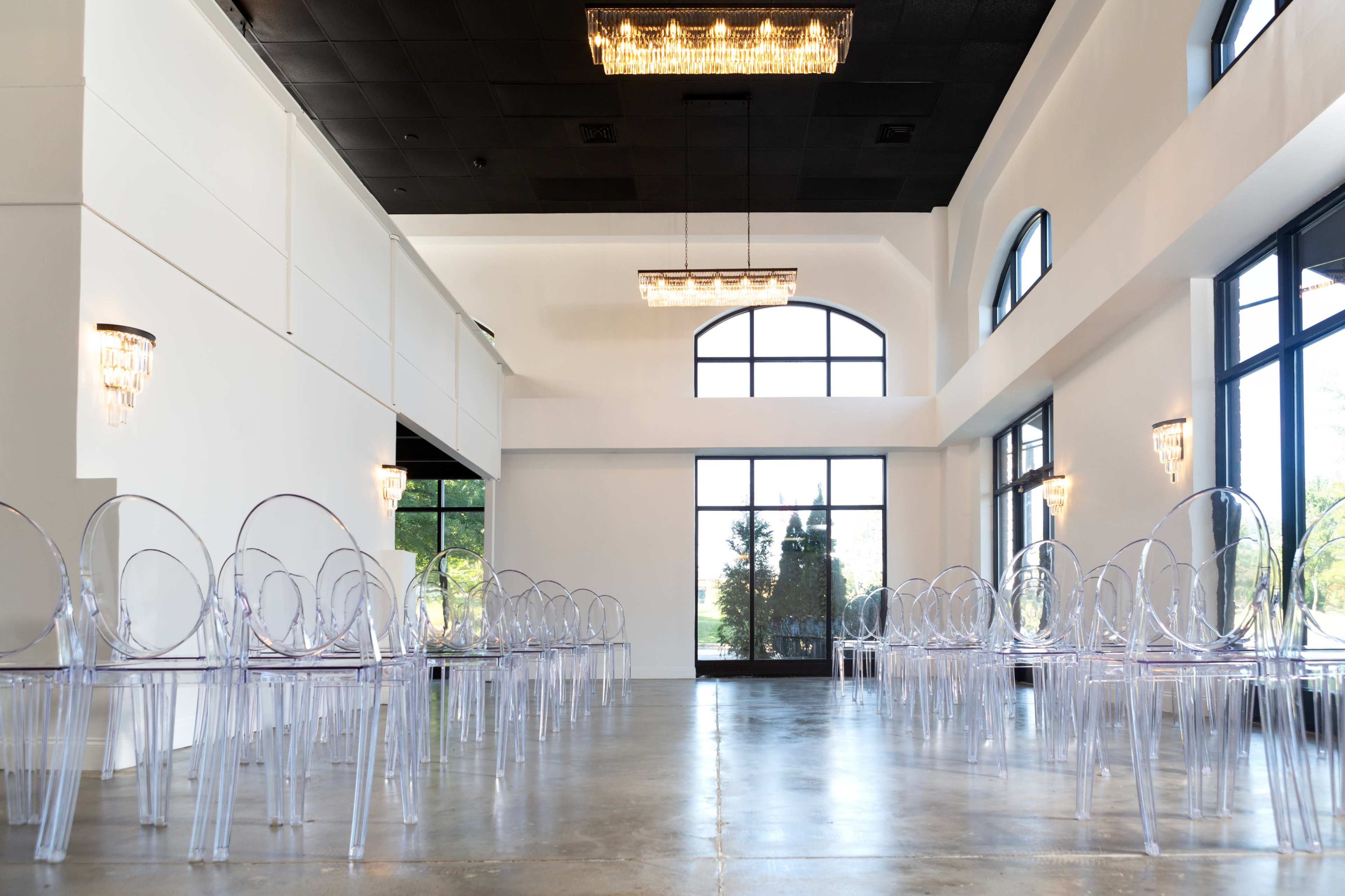 A spacious, modern indoor venue with clear acrylic chairs arranged in rows, large windows allowing natural light, and elegant light fixtures on the walls.