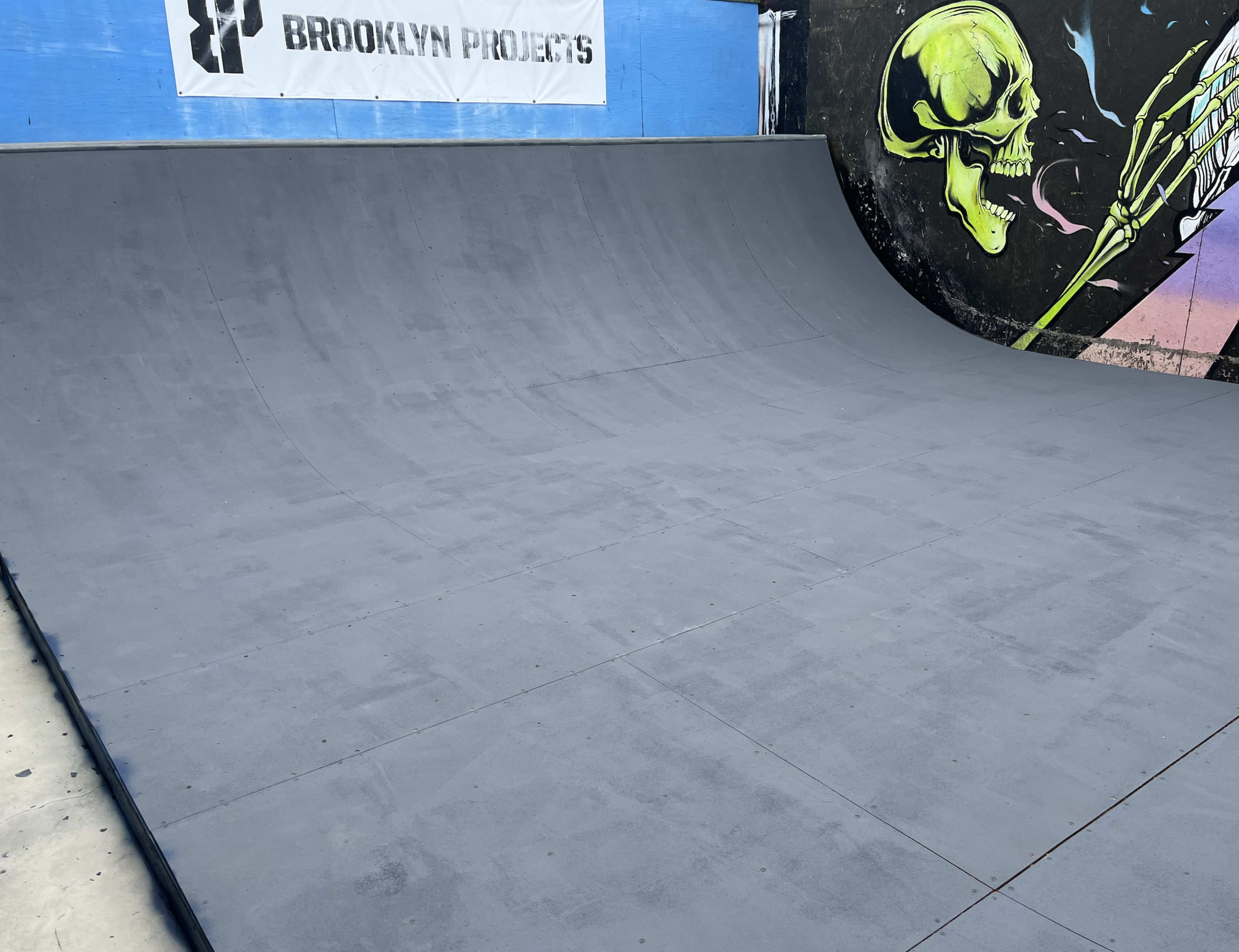 The image shows a smooth, gray skateboard ramp with a mural of a skull and checkered pattern on a nearby wall.