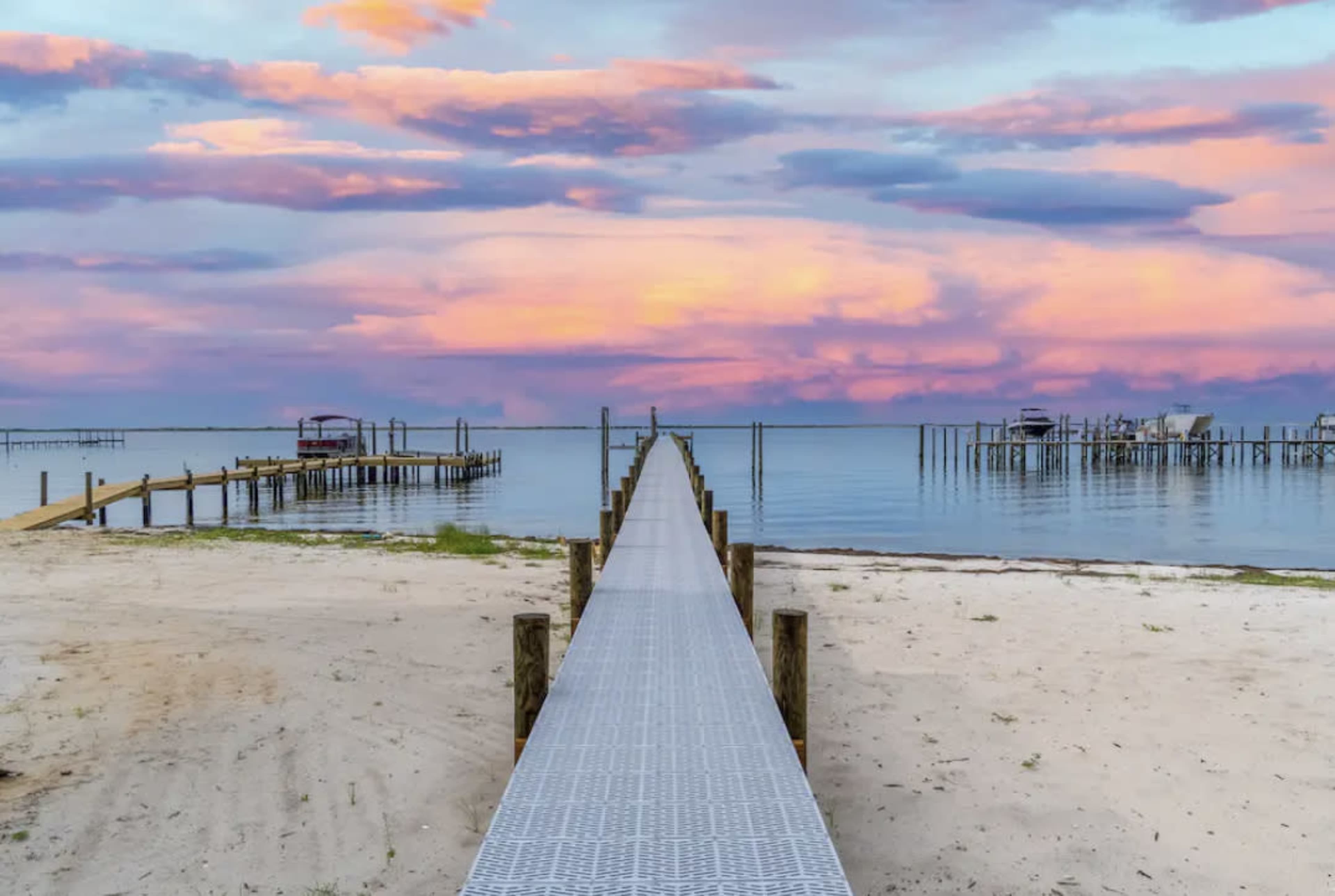 A metal dock extends over a calm body of water, leading to a horizon filled with colorful clouds at sunset.