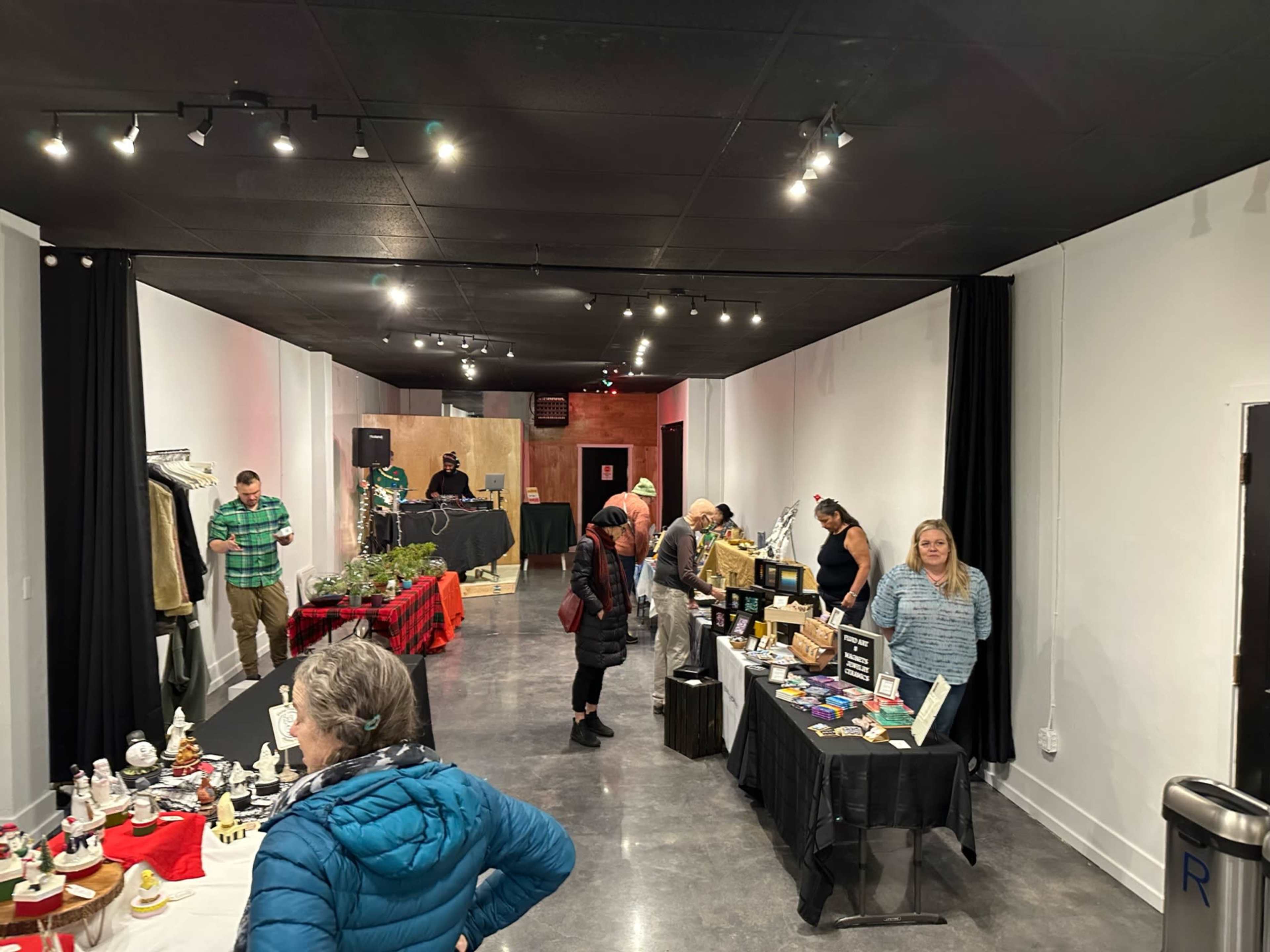 An indoor event space with vendors displaying products on tables, attendees browsing, and a DJ booth in the background.