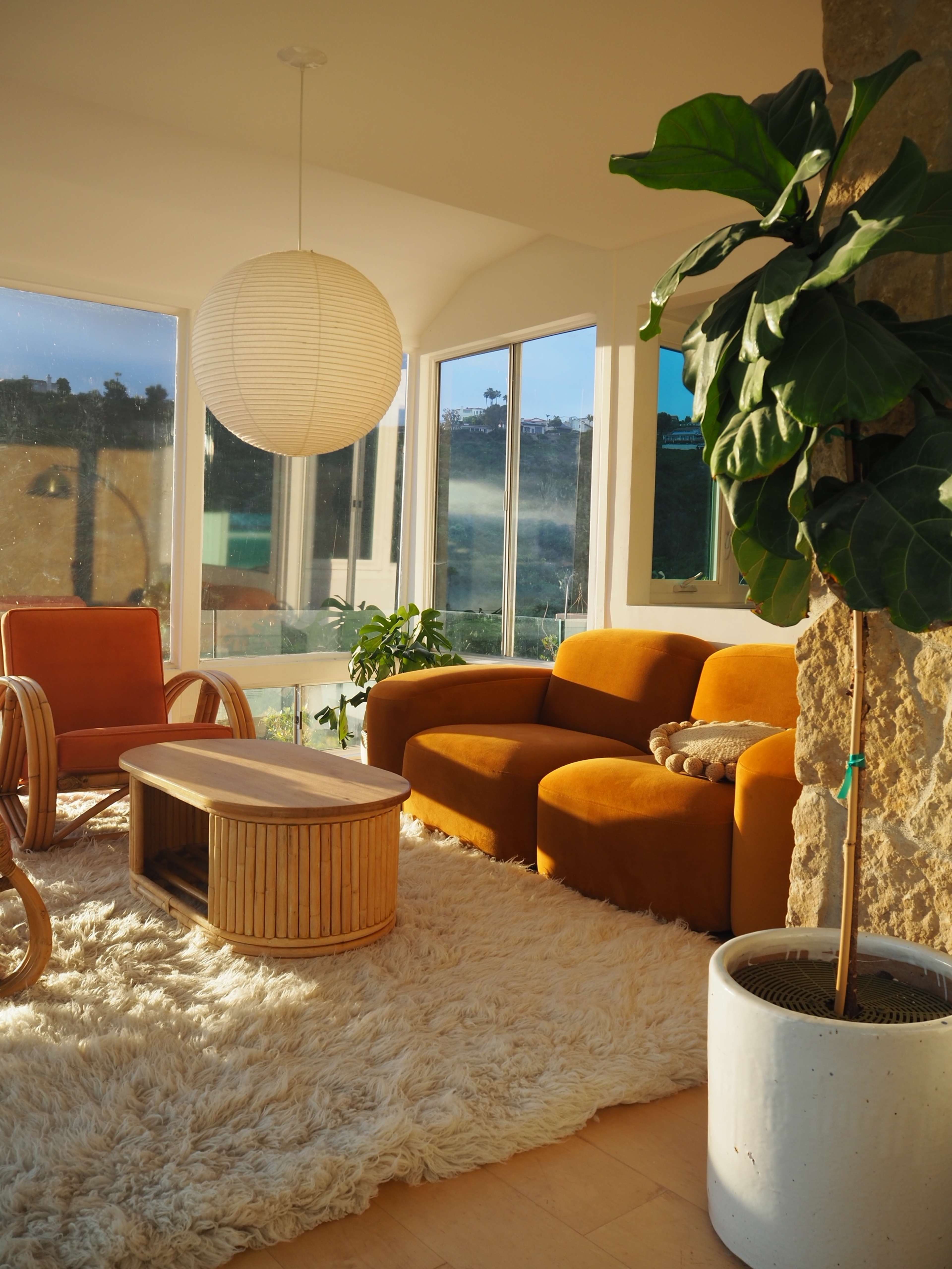 A bright living room features a wooden rocking chair, an orange sofa, a round coffee table, and a large potted plant, all arranged on a fluffy white rug.