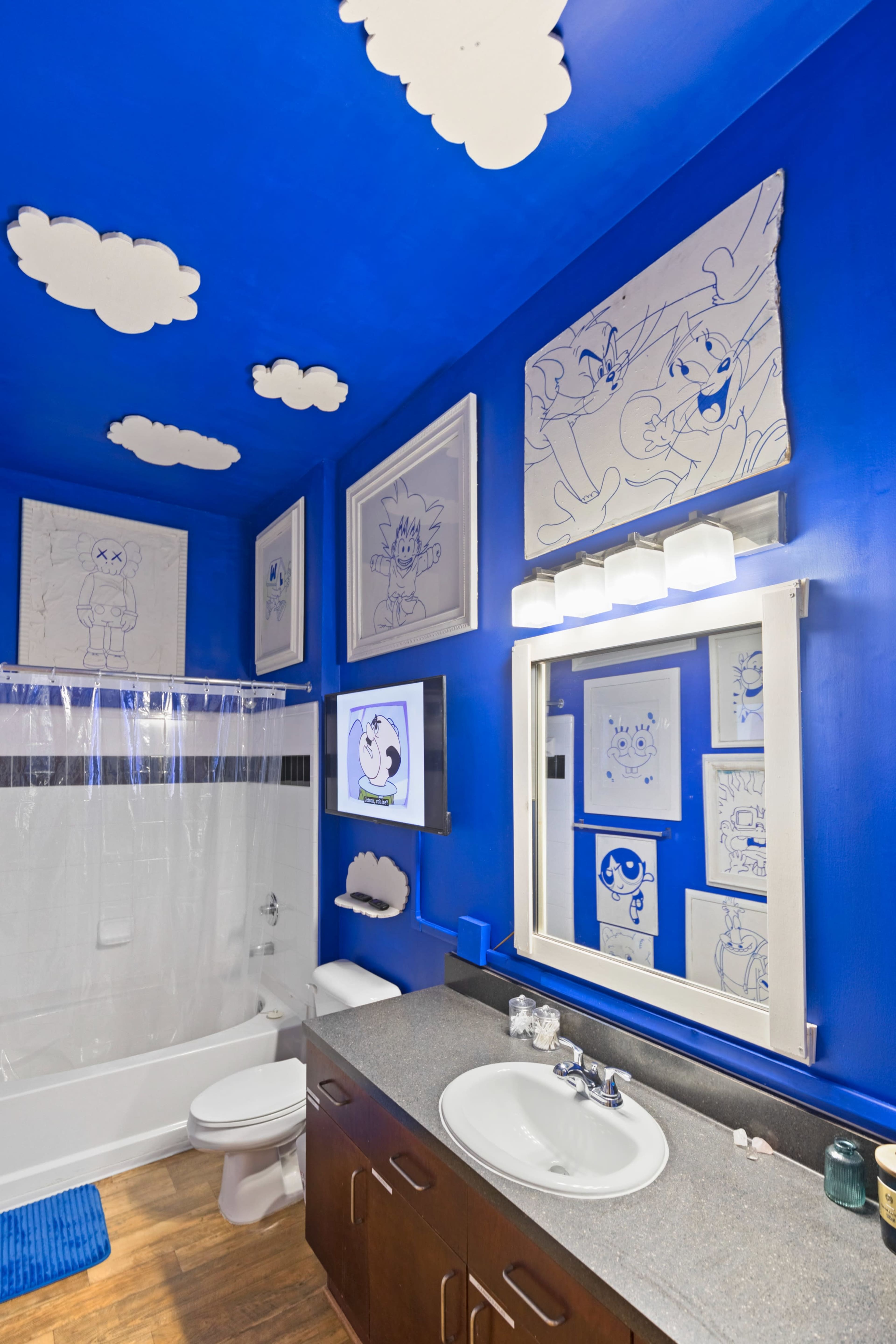 The bathroom features a blue-painted wall, a shower with a clear curtain, and framed cartoon illustrations on the walls.