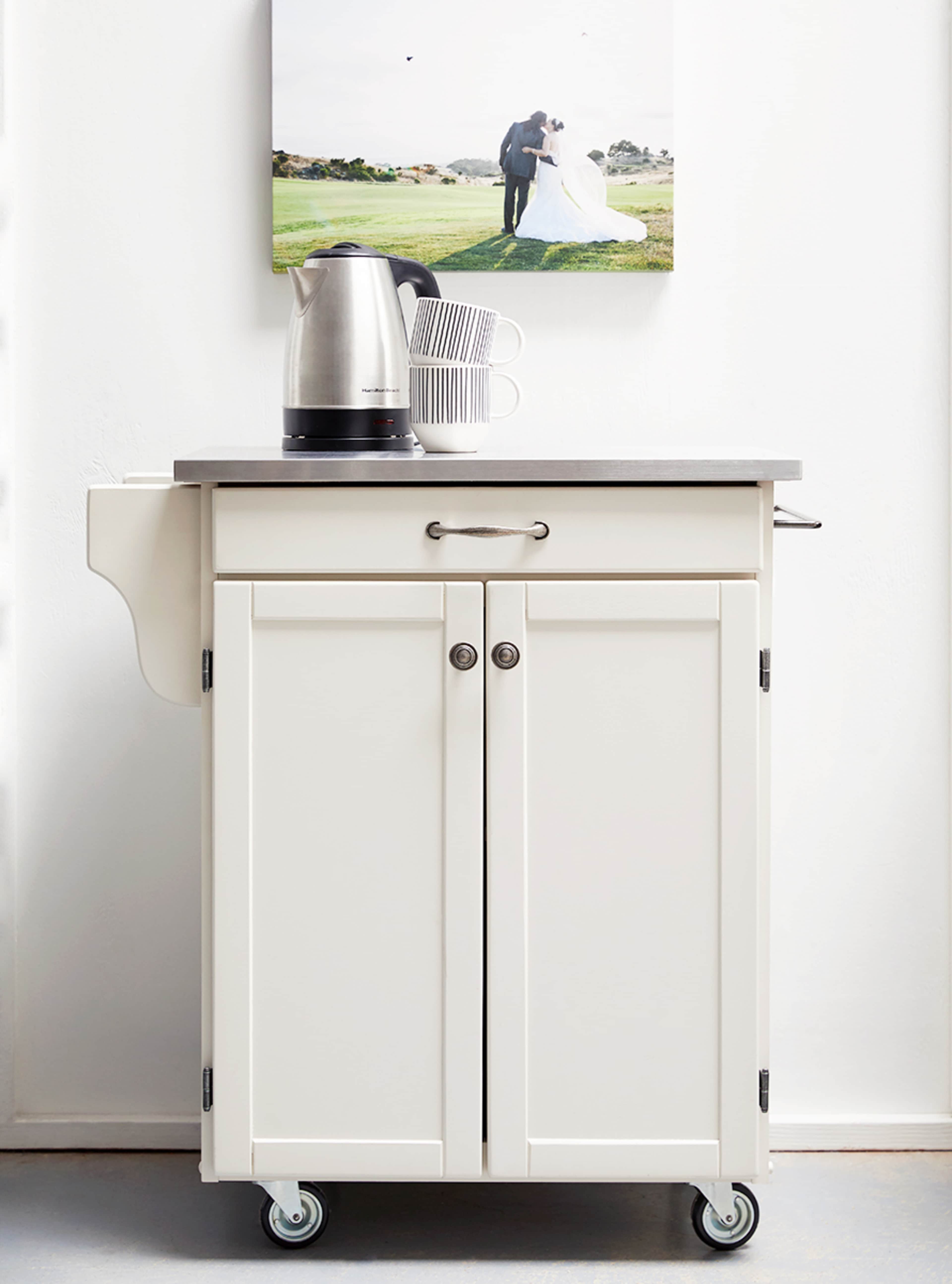 A white kitchen cart with a stainless steel kettle and stacked dishes on a gray countertop, positioned against a white wall featuring a framed photograph.
