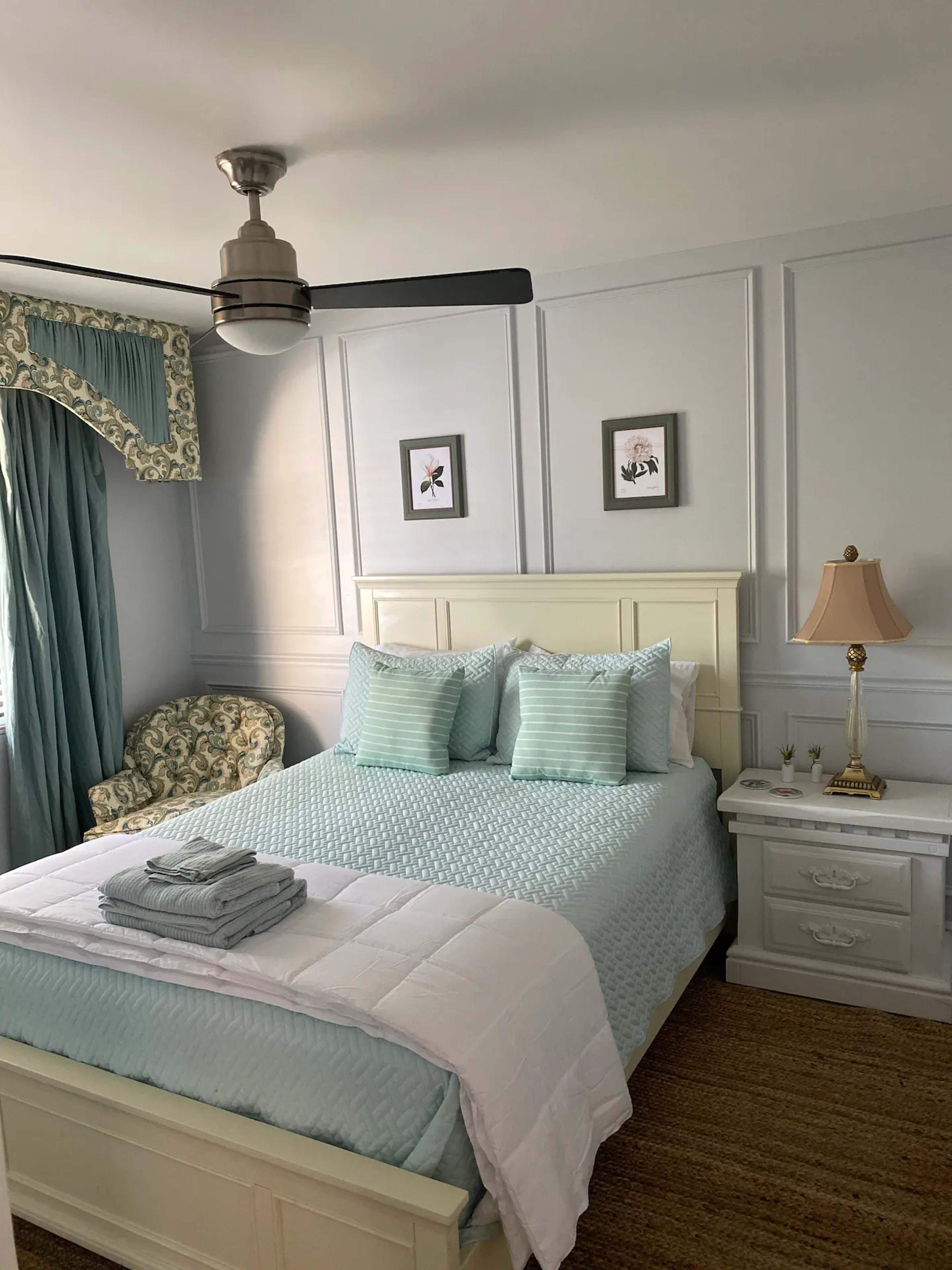 The image depicts a neatly arranged bedroom featuring a light blue bedspread, decorative pillows, a bedside lamp, and a patterned armchair near a window.