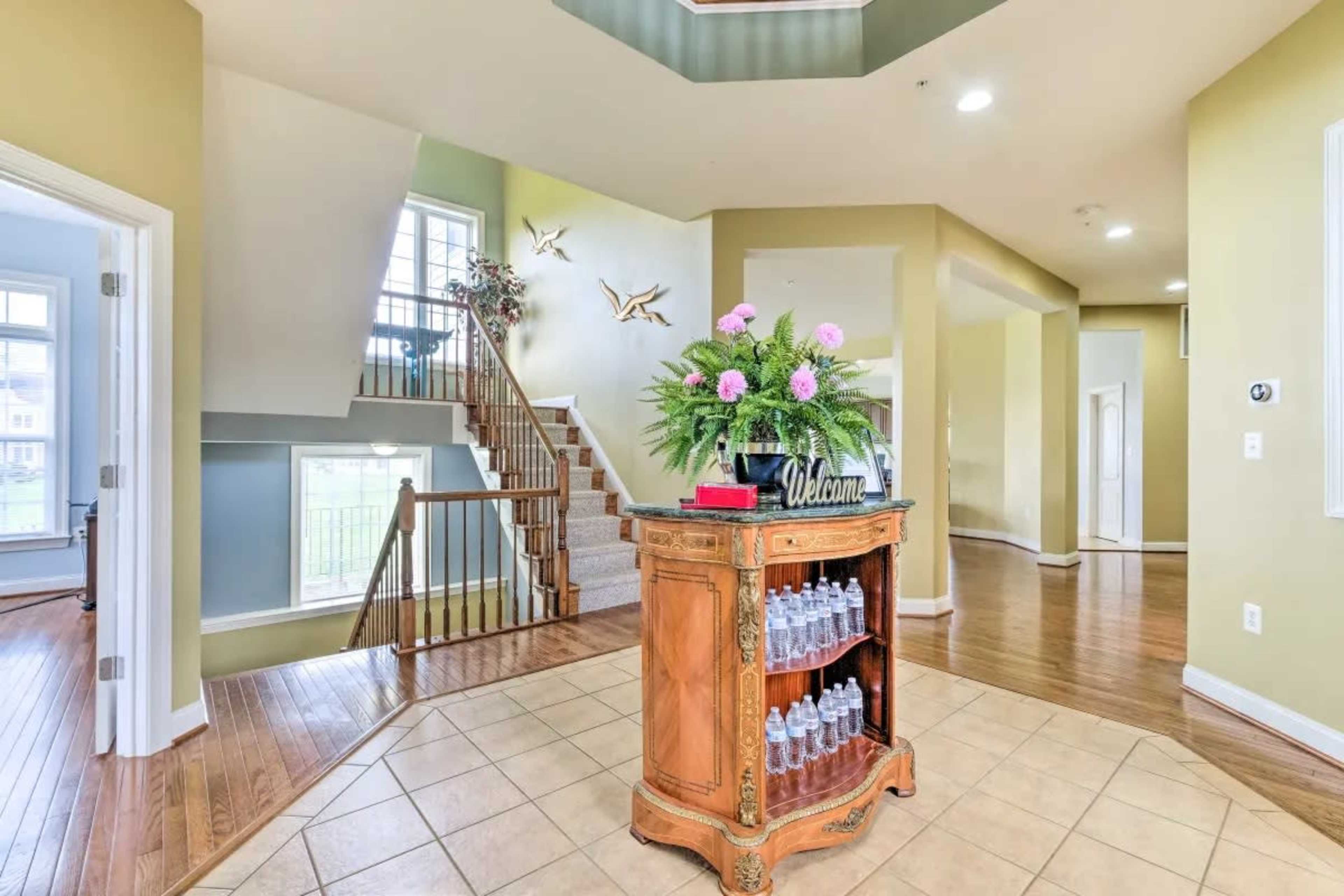 A spacious entryway with a wooden staircase, a decorative console table with water bottles, and large windows allowing natural light.