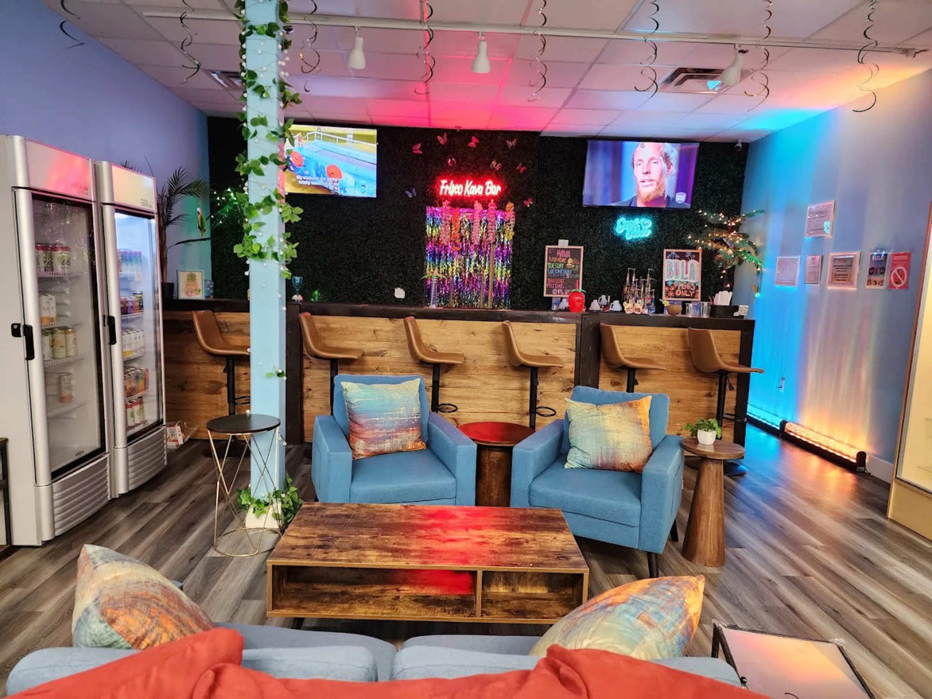 The image shows a modern lounge area featuring a bar with high stools, various drinks displayed in a refrigerator, and a cozy seating arrangement with blue chairs and a wooden coffee table, all adorned with decorative lighting.