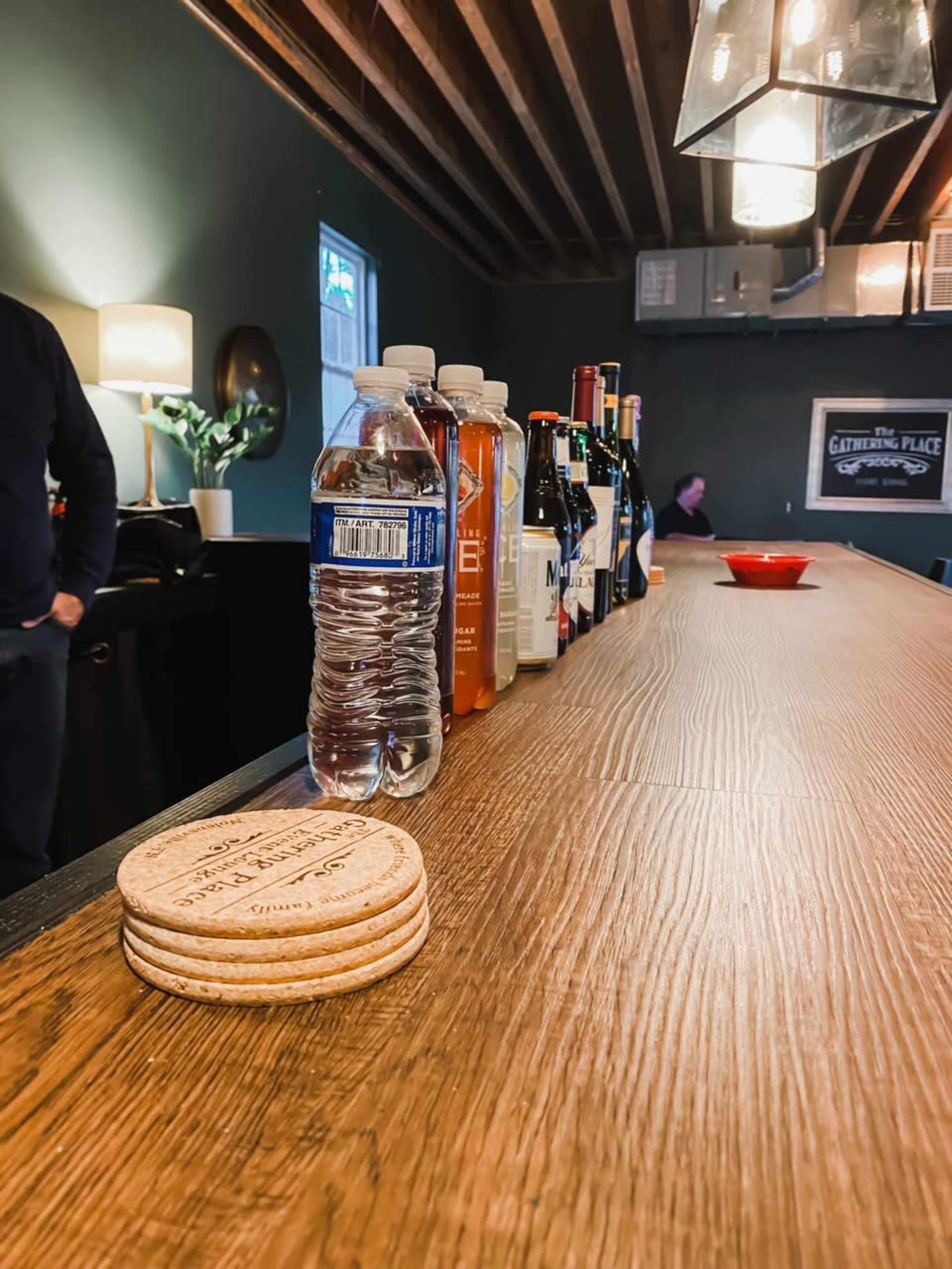 A wooden bar countertop displays a lineup of various beverages, including a bottle of water and several liquor bottles, alongside coasters.