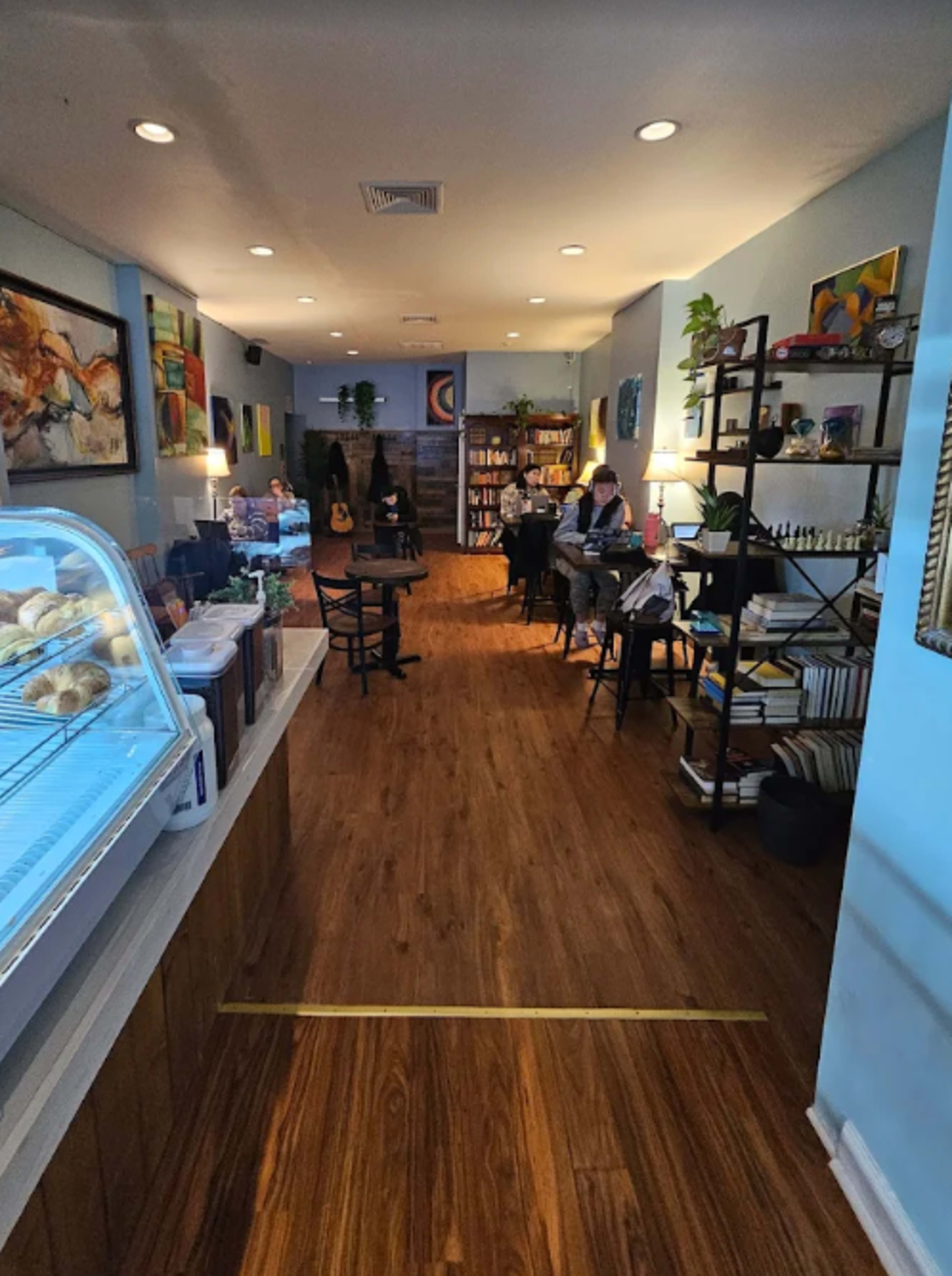 The image shows a cozy café interior with wooden flooring, tables arranged along the sides, and a display case for pastries at the front.