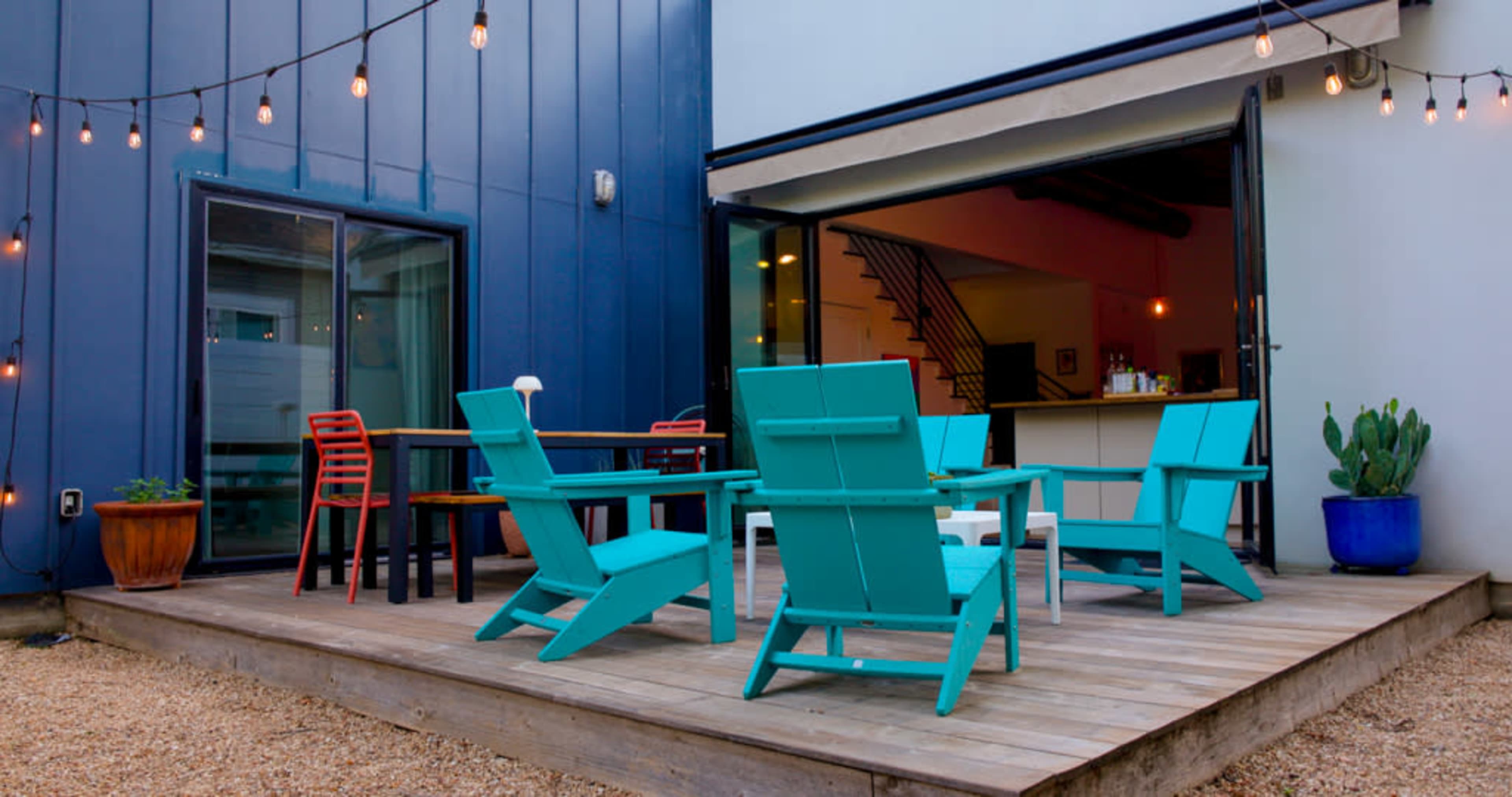 The image shows a wooden deck with turquoise outdoor chairs, a table, and string lights, leading to an open door that reveals an interior space.