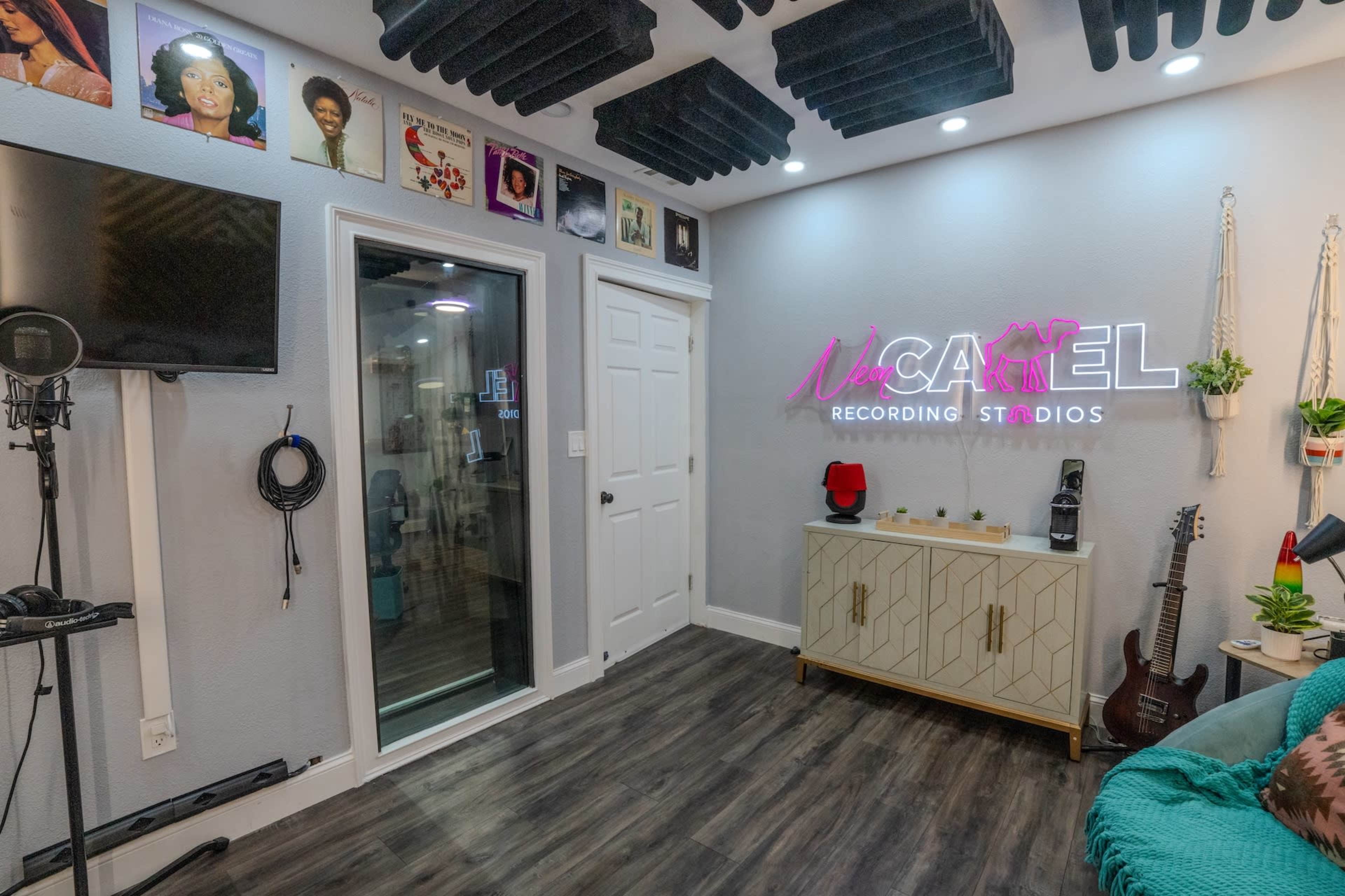 The image shows a modern recording studio interior featuring a neon sign, various framed portraits on the walls, and soundproofing panels on the ceiling.