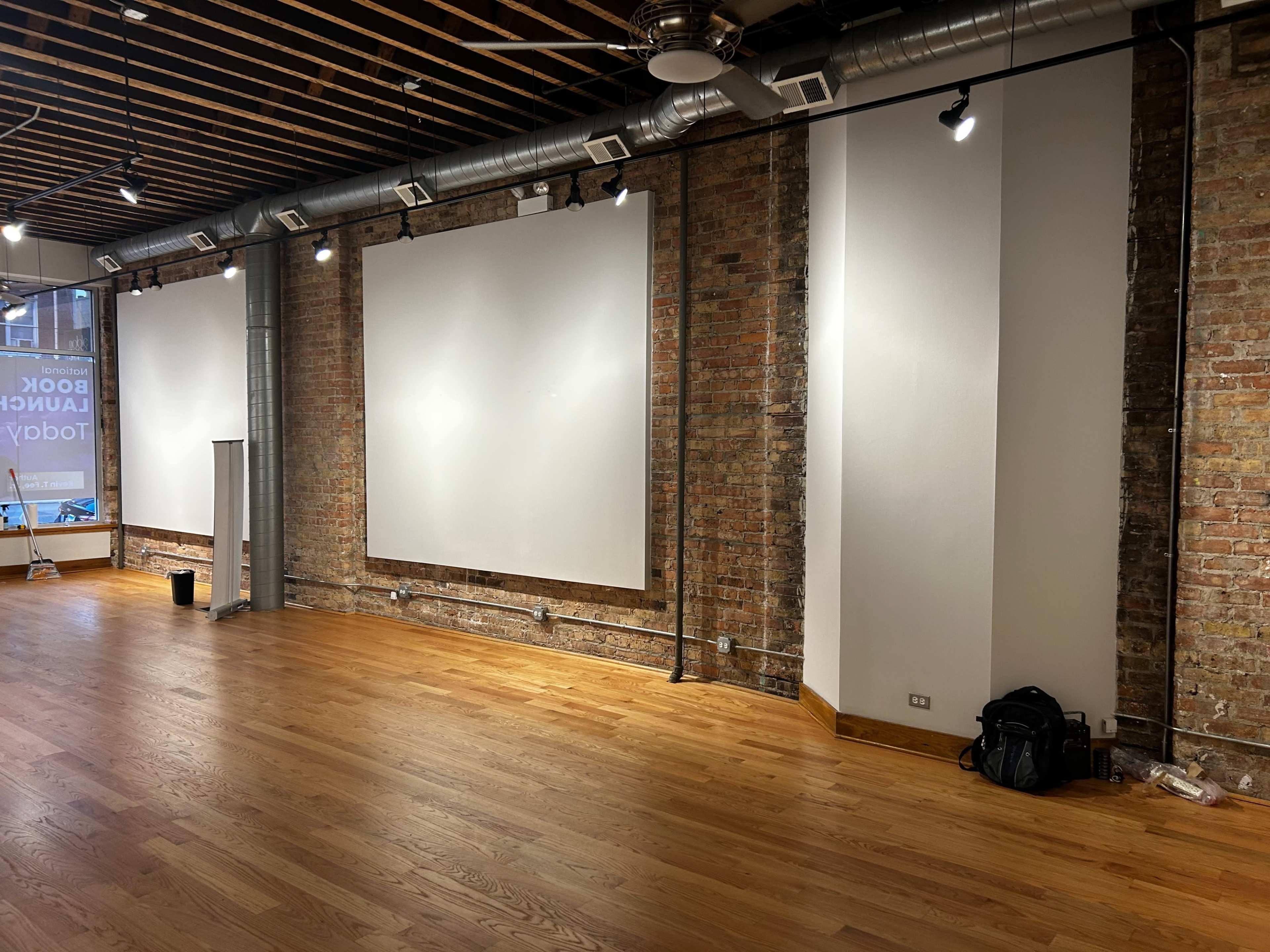 The image shows an empty, well-lit room with exposed brick walls, wooden flooring, and two large blank canvases mounted on the wall.