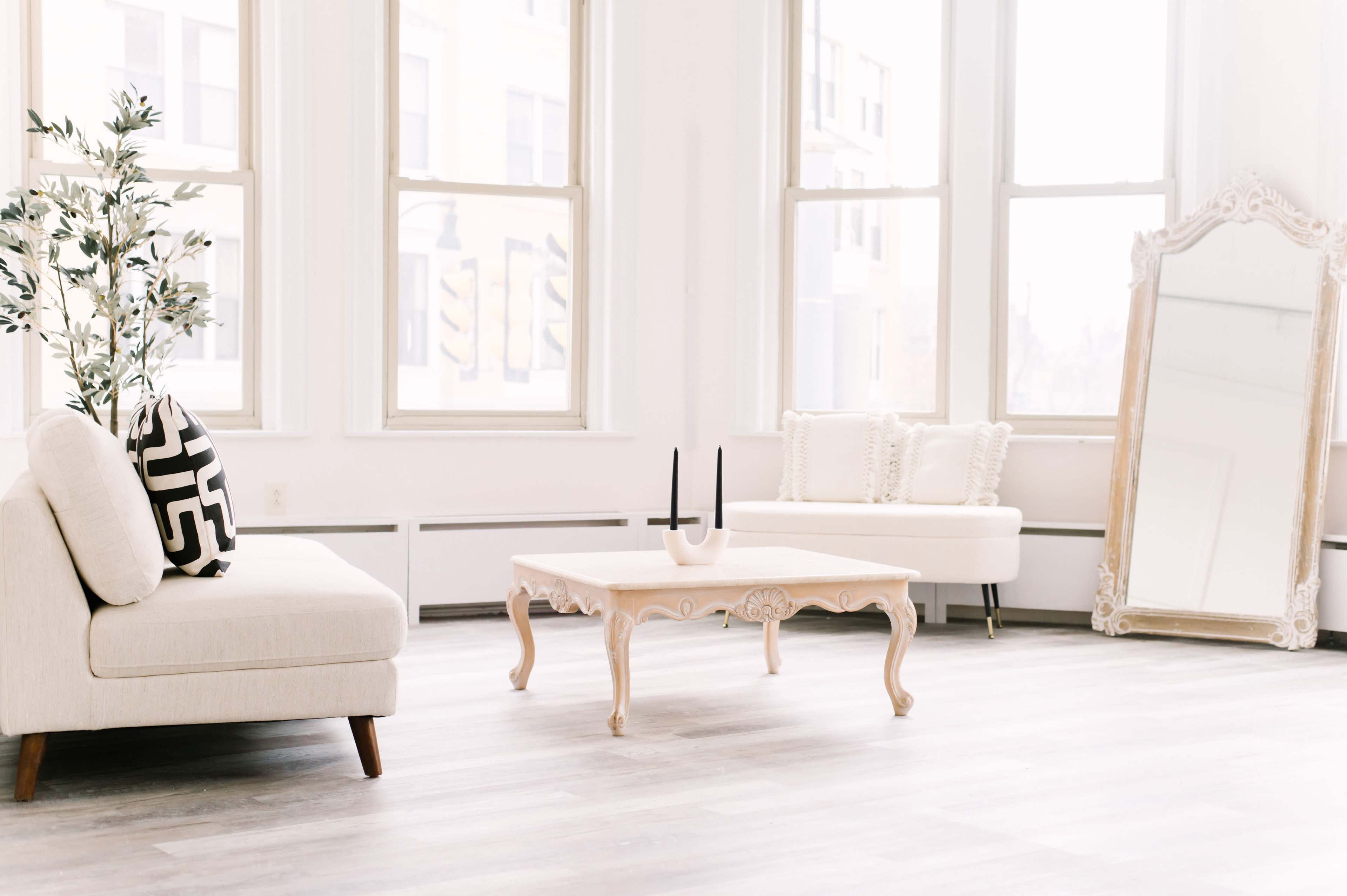 A bright, modern living space with a light-colored sofa, a decorative coffee table, and a large mirror against a wall with windows.