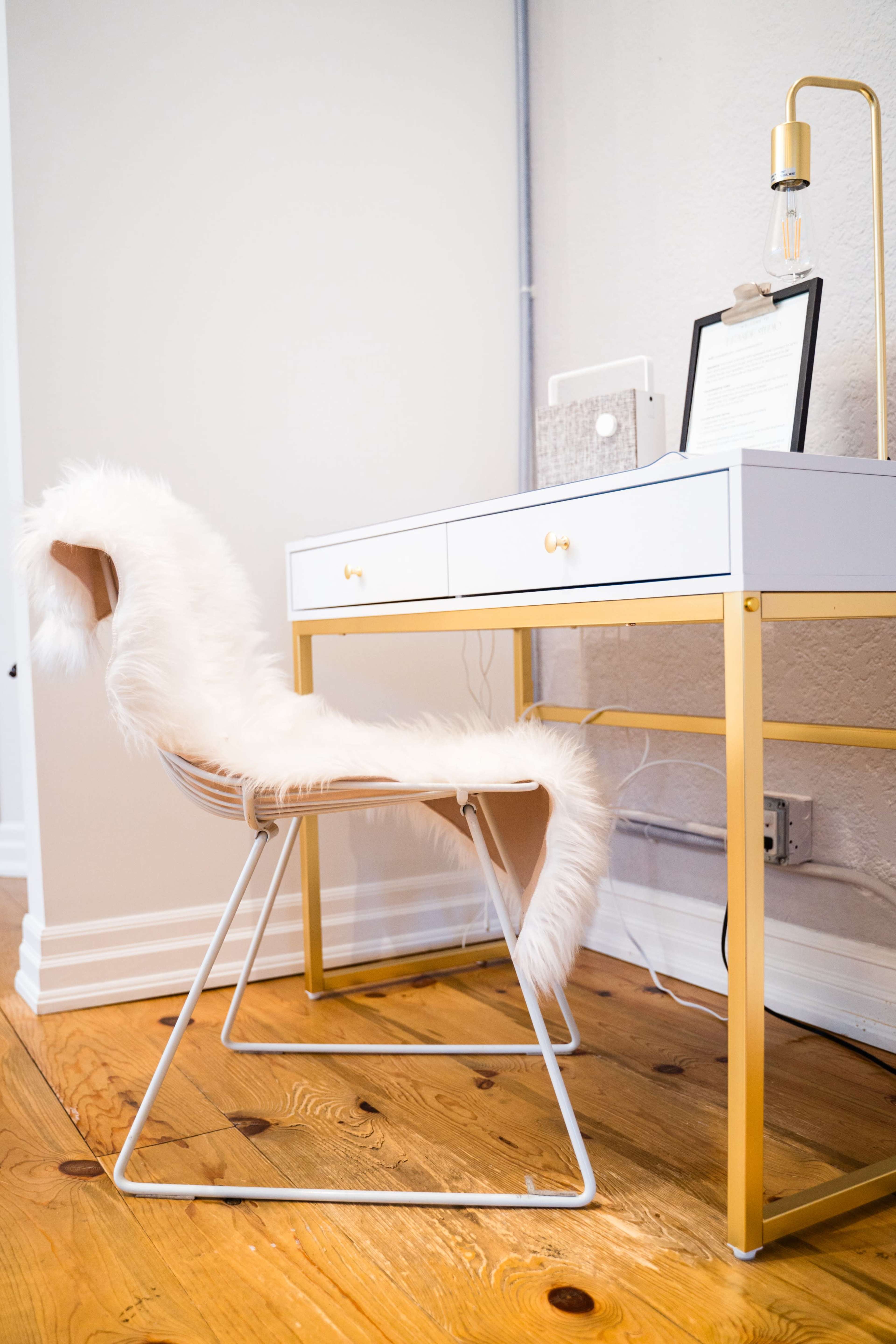 A modern desk with two drawers is positioned beside a chair draped in a white faux fur, set on a wooden floor.