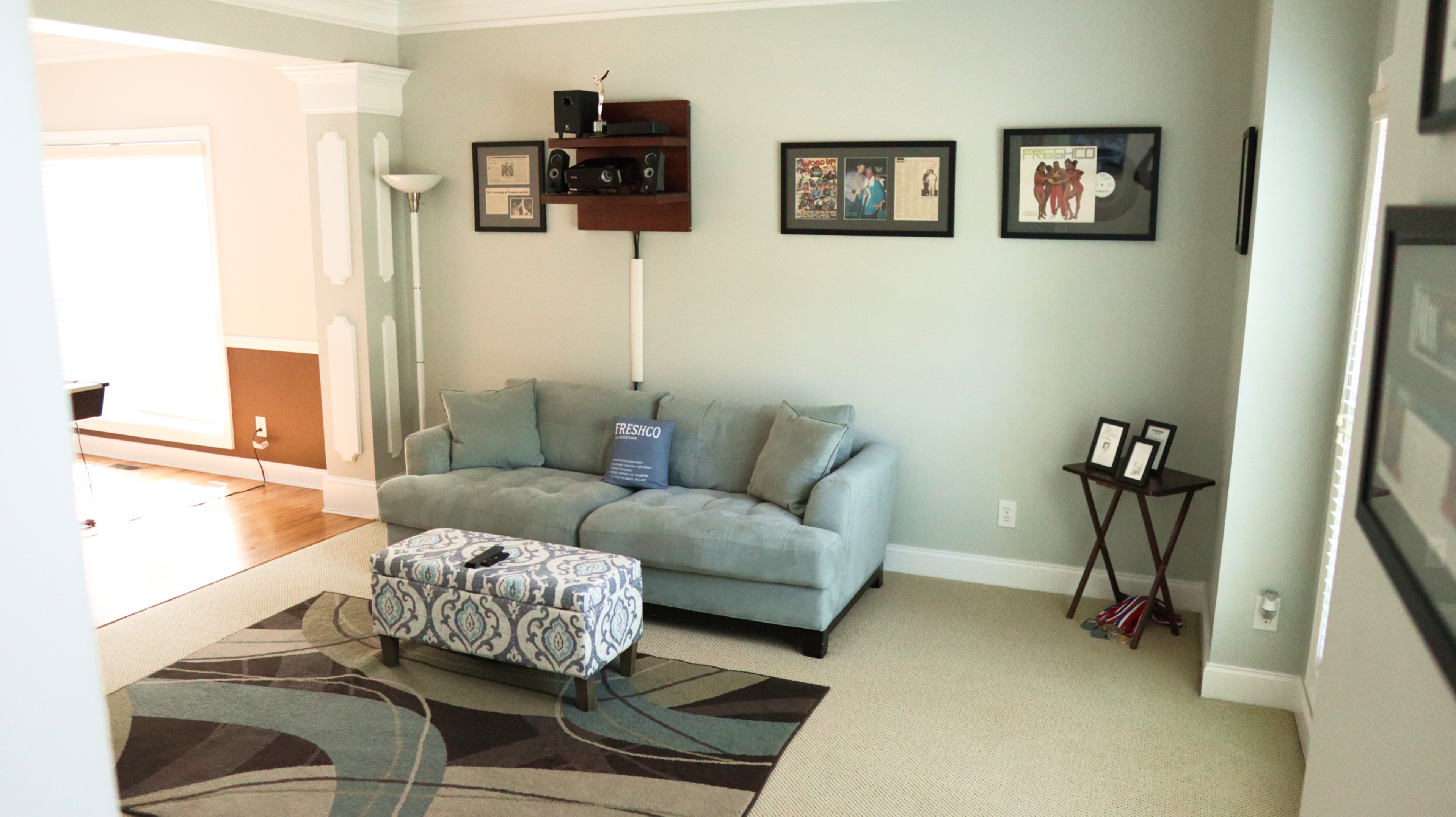 A well-lit living room with a light green wall, a gray couch, a patterned ottoman, and framed pictures on the walls.