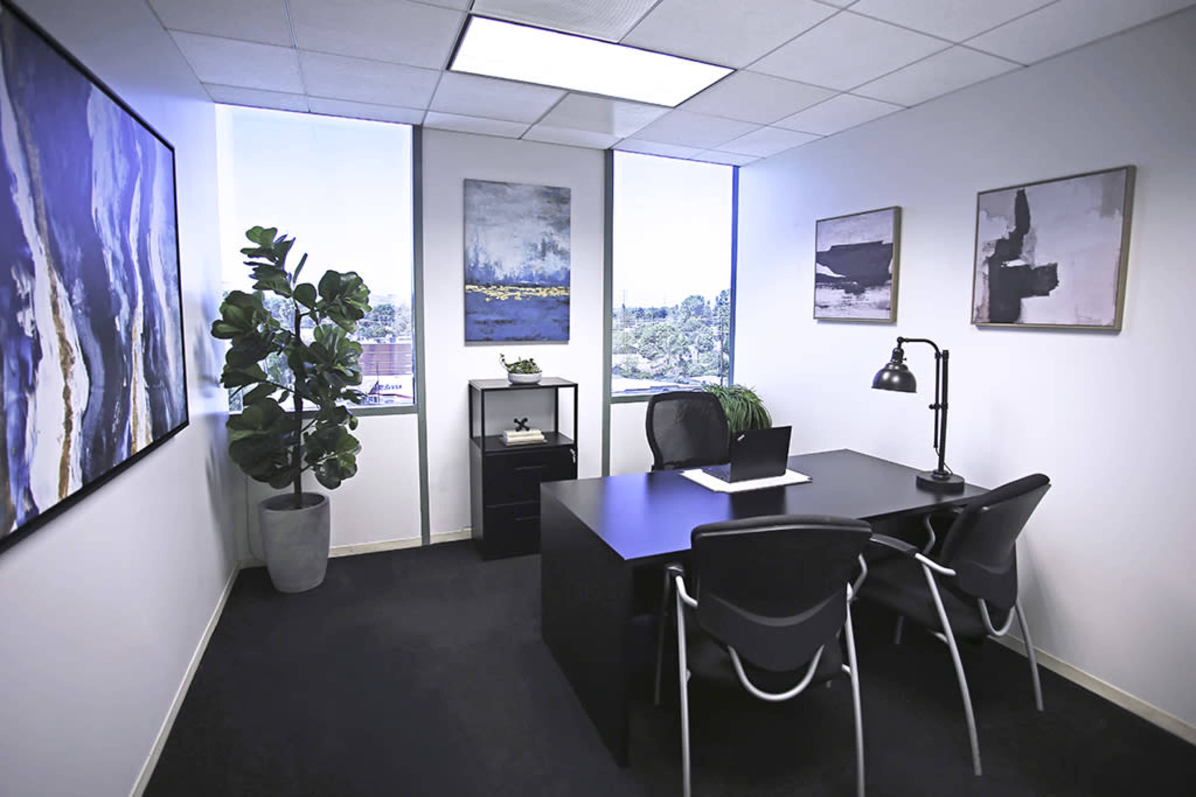 A small office space featuring a black desk with two chairs, framed artwork on the walls, a potted plant, and a window that offers a view of the outdoors.