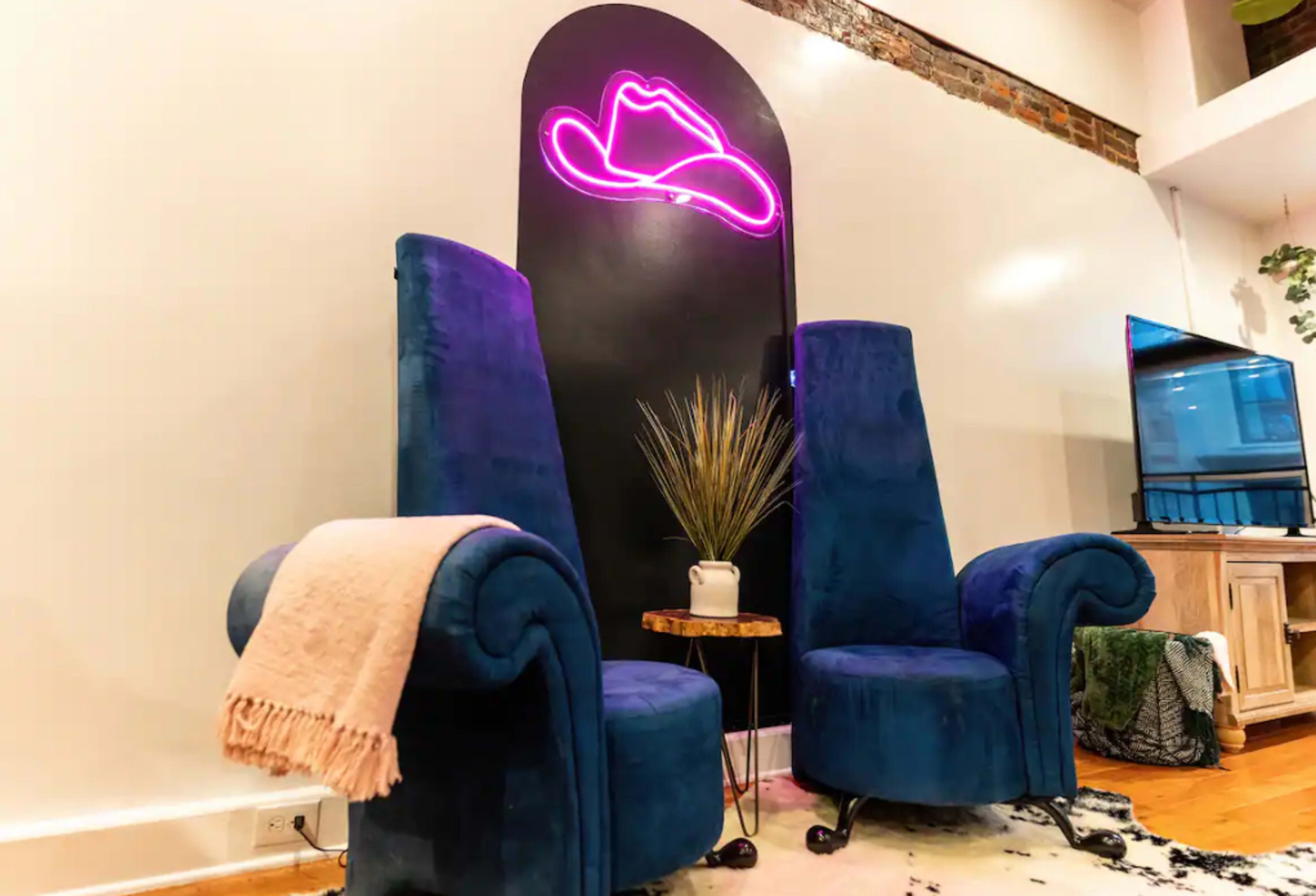 A set of two blue velvet chairs is positioned next to a small wooden table with a plant, set against a wall featuring a pink neon cowboy hat sign.