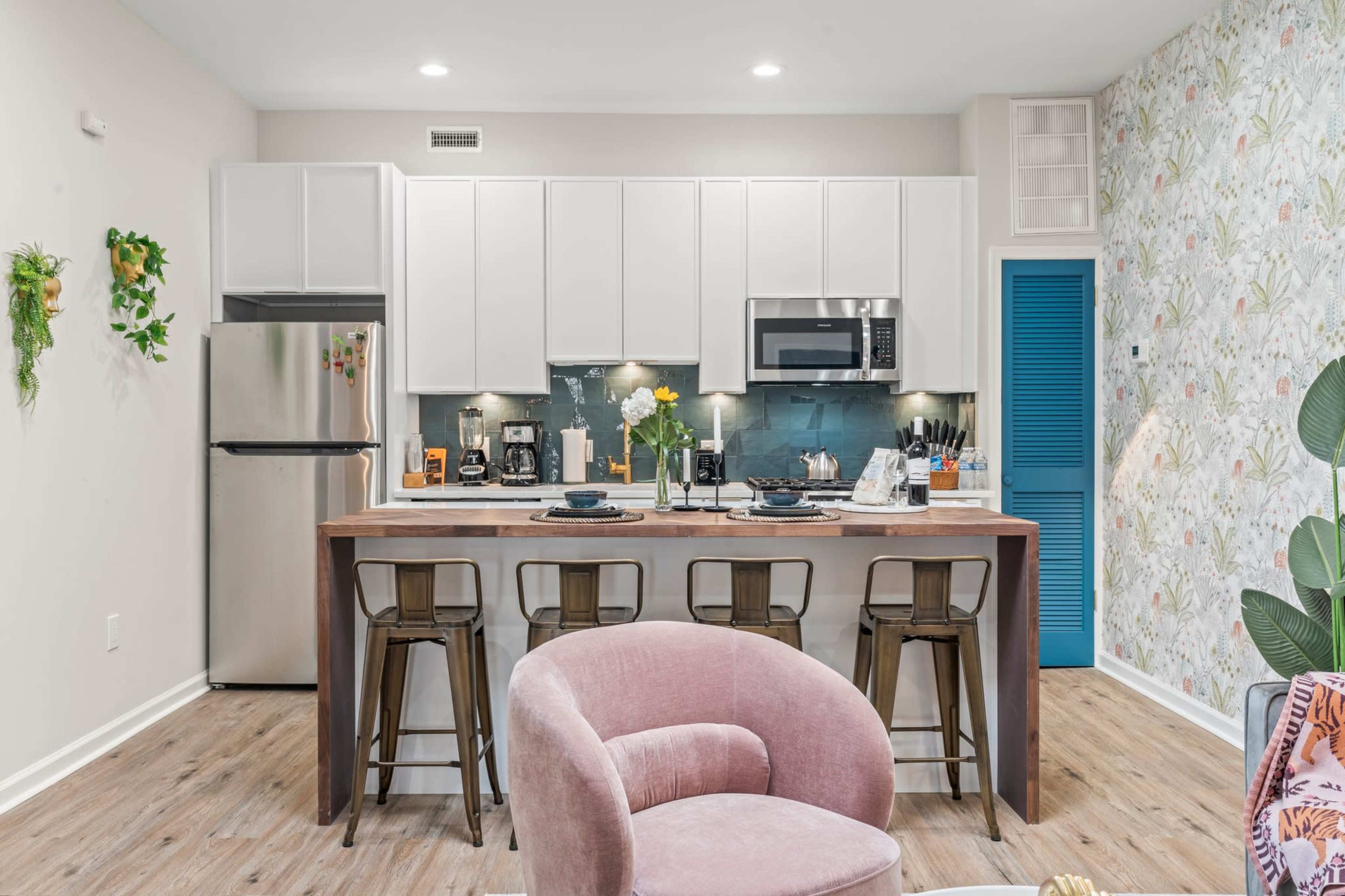 The image shows a modern kitchen with white cabinets, stainless steel appliances, and a wooden island with bar stools, featuring decorative wallpaper and plants.