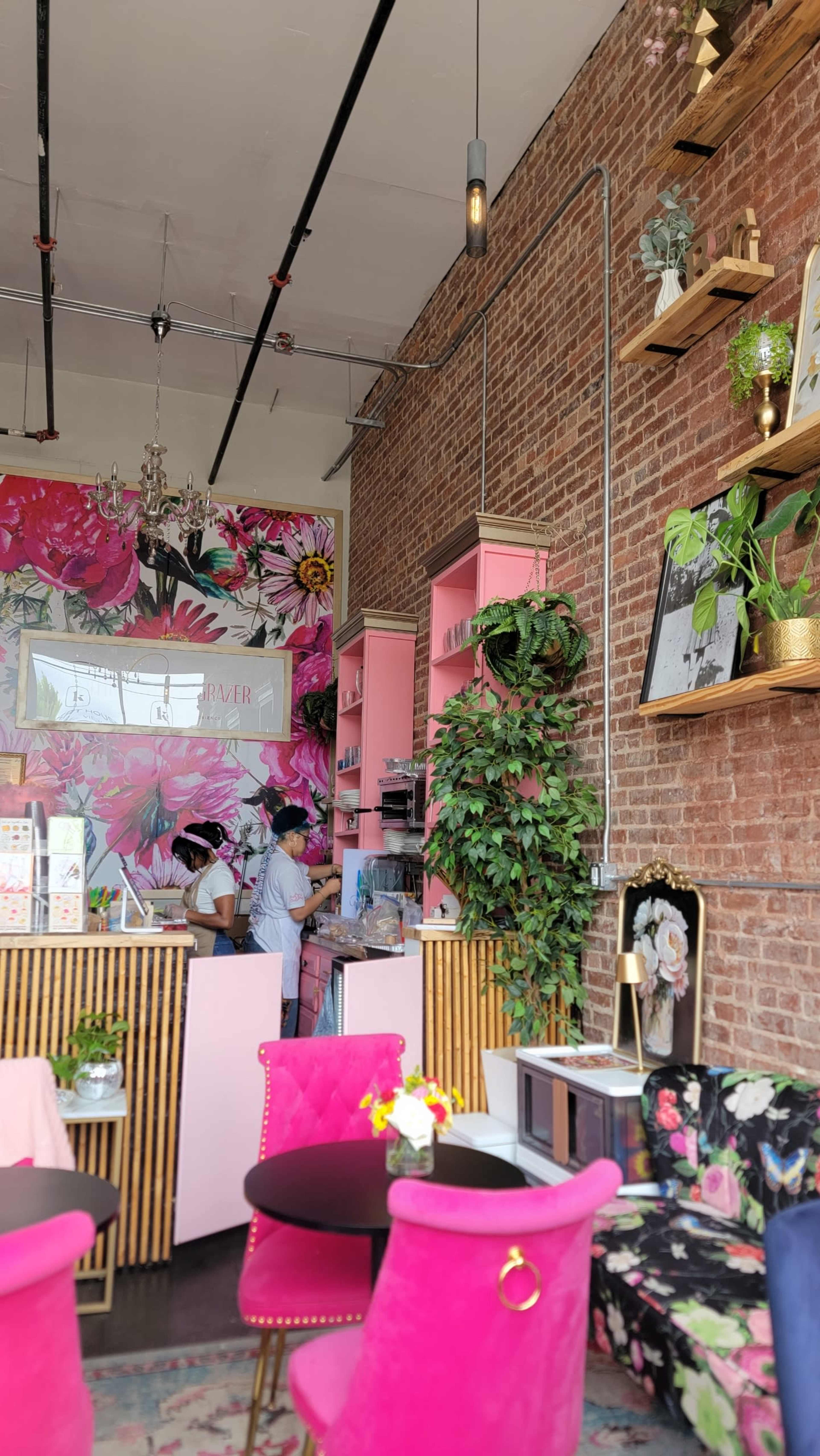 The image shows a vibrant cafe interior with pink furniture, a floral mural on the wall, and a staff member preparing drinks behind a counter.