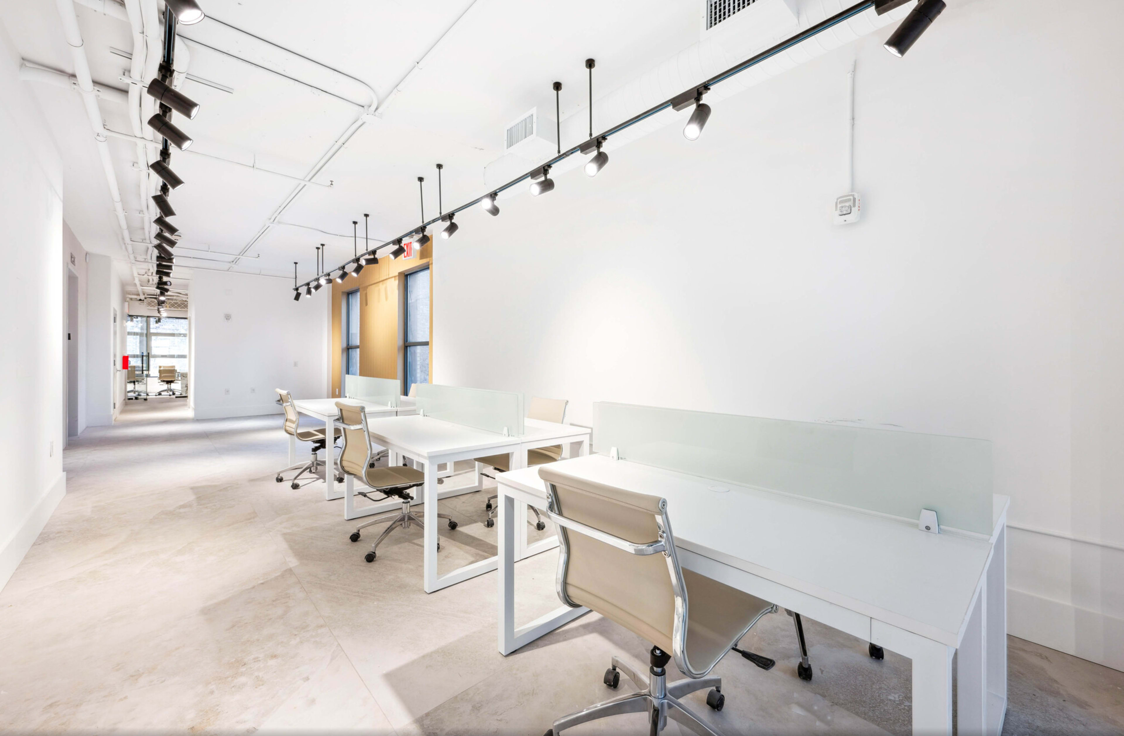 The image shows a bright, modern office space with several white desks and ergonomic chairs arranged along a long, narrow corridor.