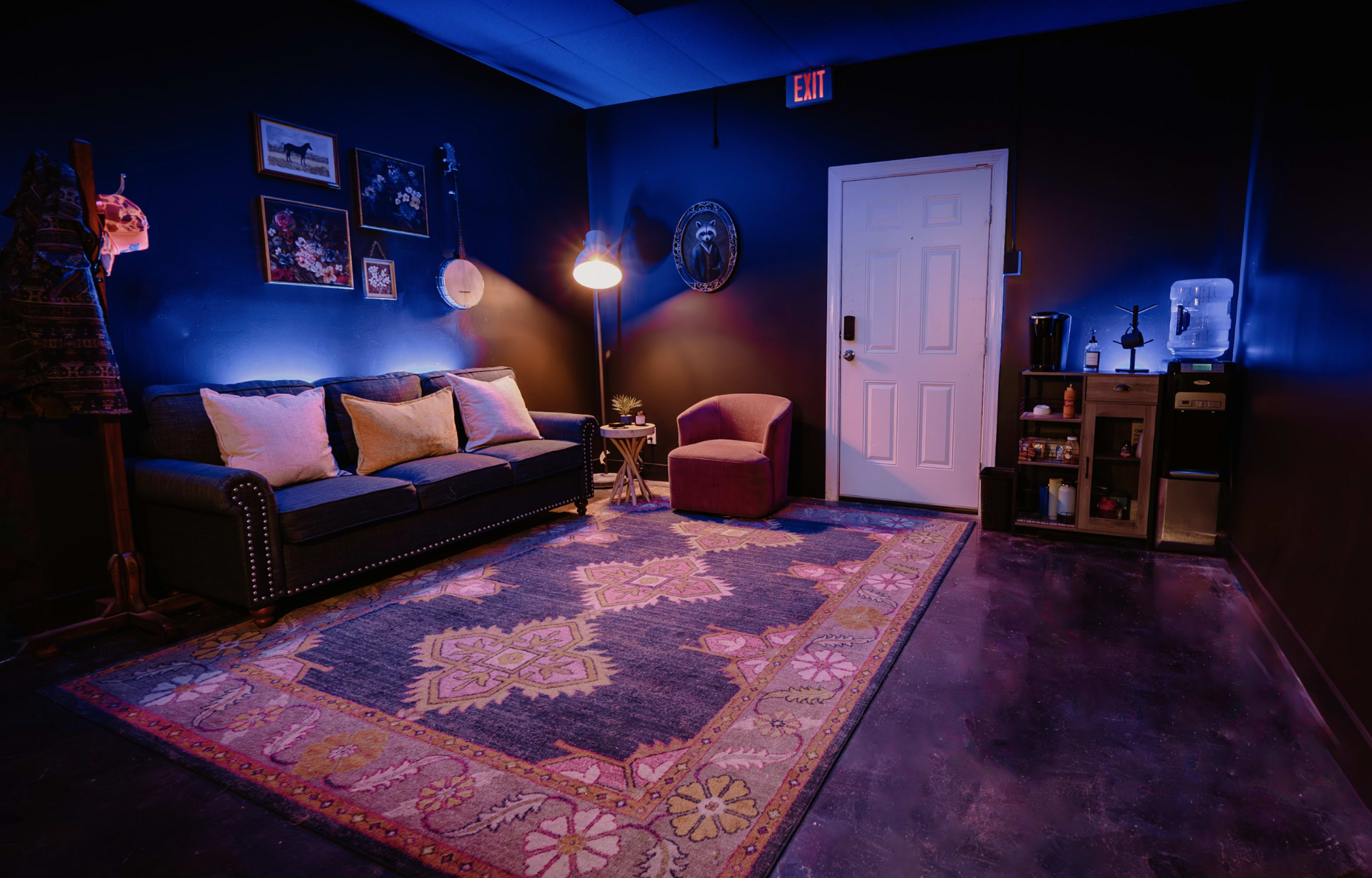 The image shows a small room featuring a dark-painted wall, a sofa with cushions, an armchair, a decorative rug, and a door with a light fixture.