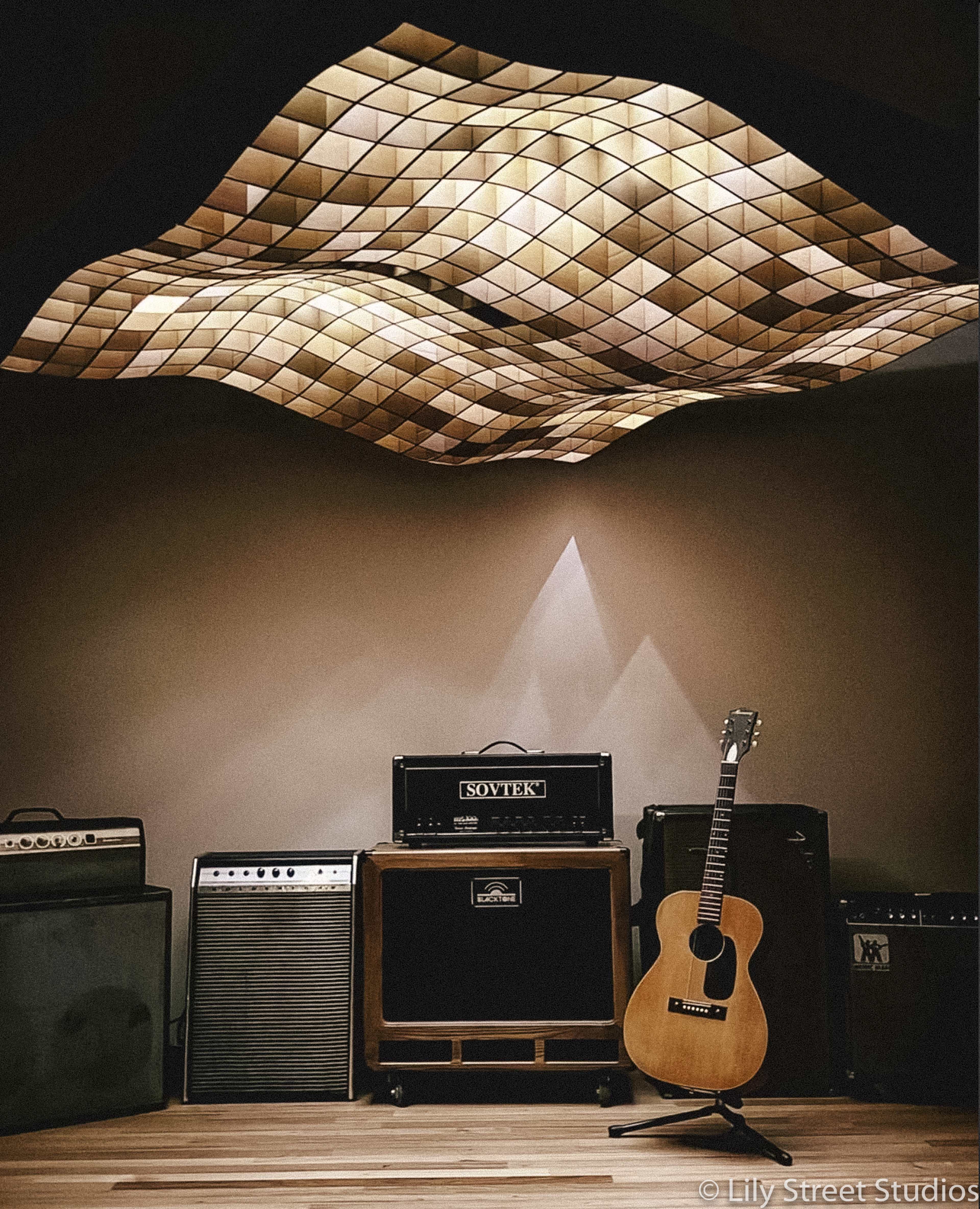 Several amplifiers and a wooden acoustic guitar against a wall, illuminated by soft lighting from a uniquely shaped ceiling fixture.