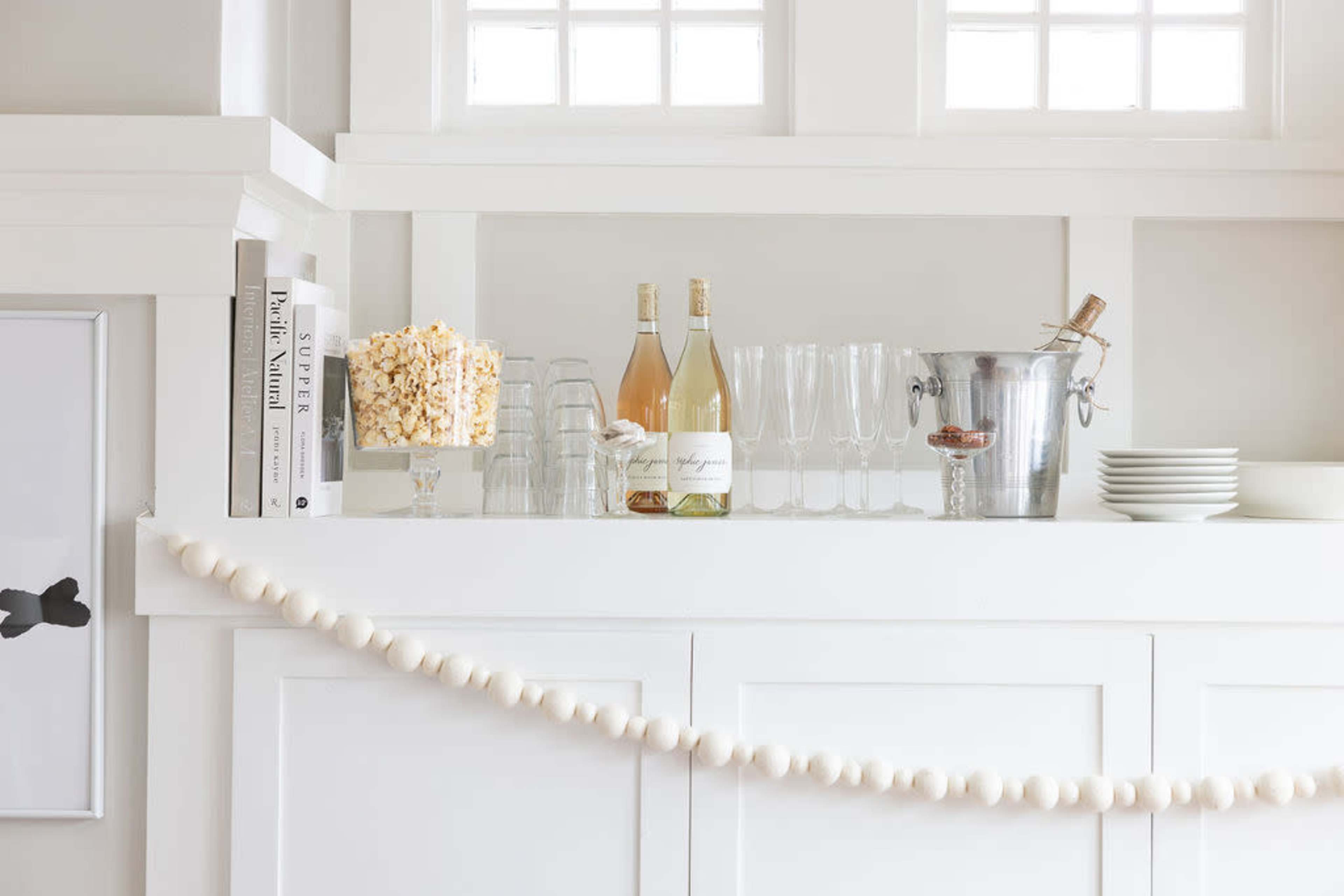 A white shelf displays glasses, a wine bottle, popcorn in a clear container, and a metal bucket, with a decorative string of beads hanging below.