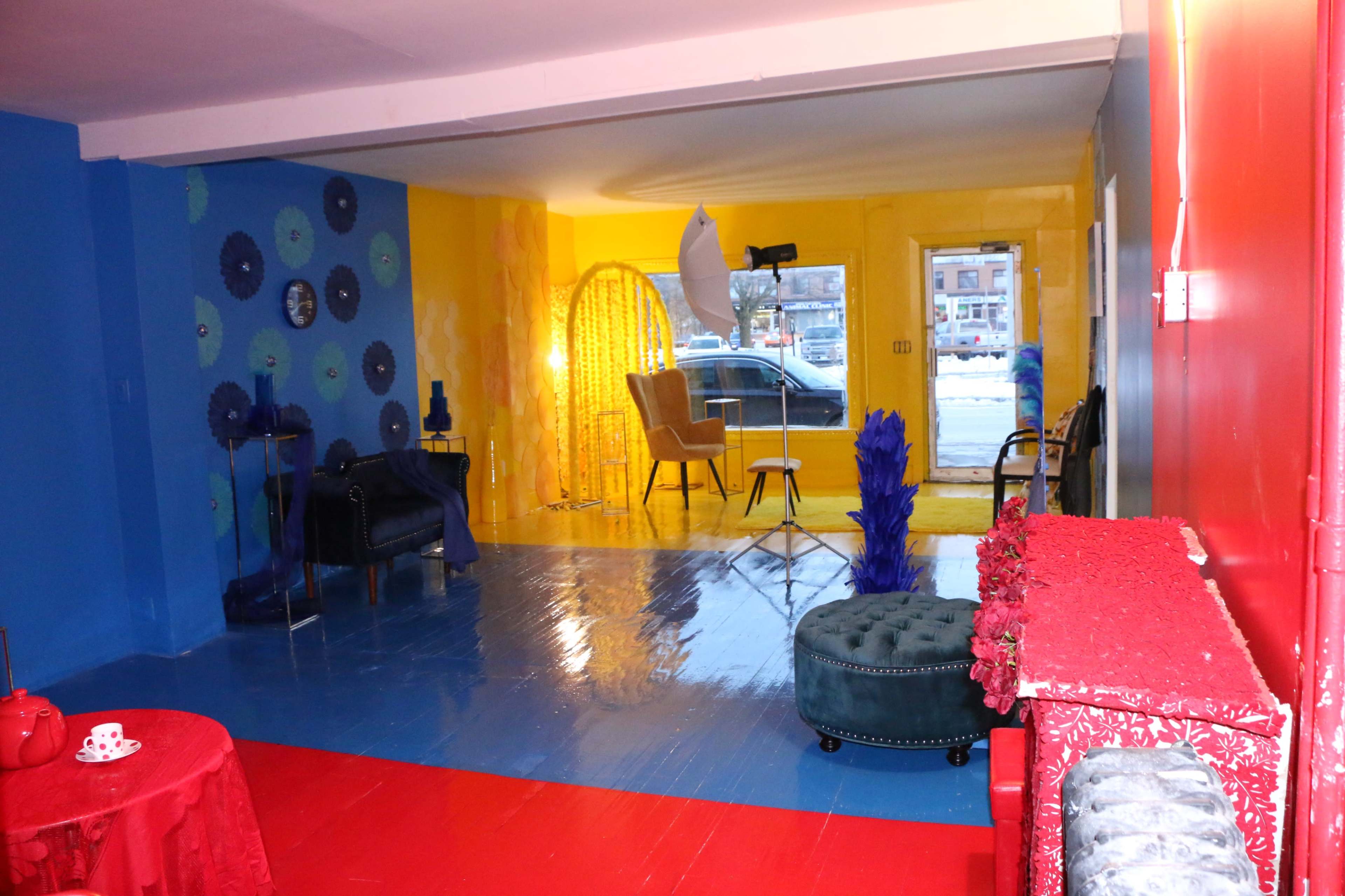 A brightly colored room with blue and yellow walls, a red floor, and furnishings including a sofa, chairs, and a light setup for photography.