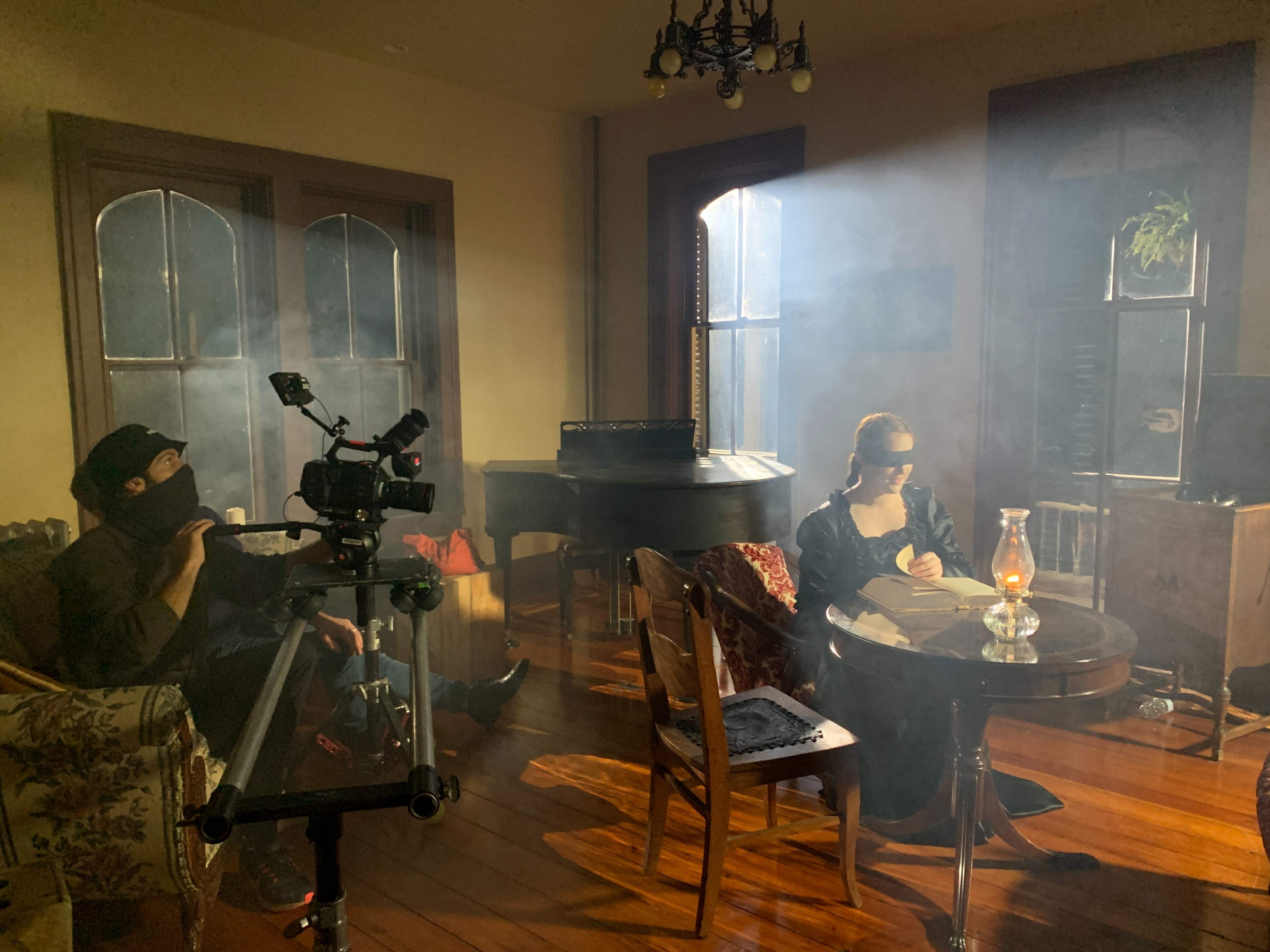 A person wearing a blindfold sits at a table with a lantern, while another person operates a camera in a softly lit room filled with smoke.