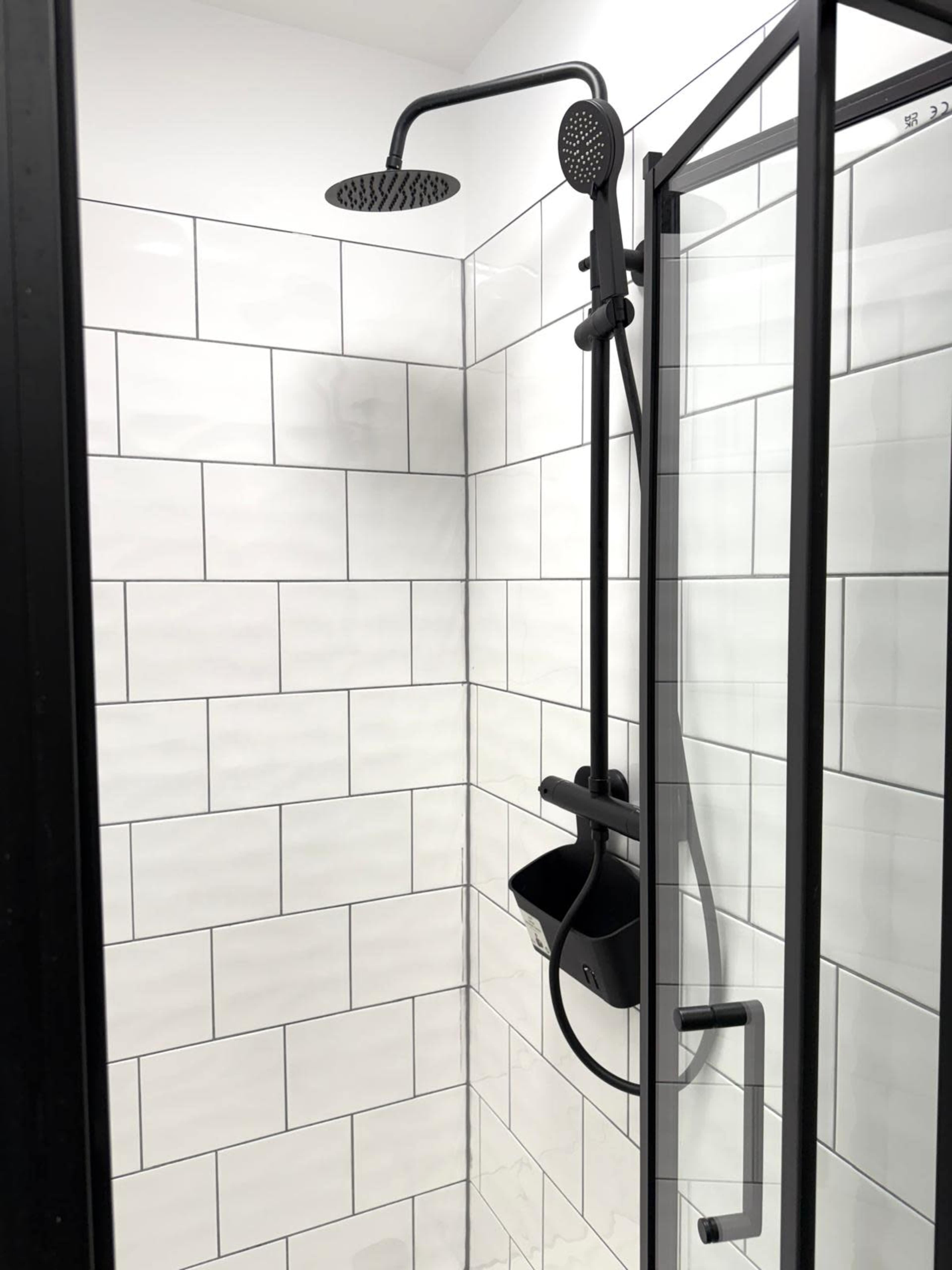 The image shows a modern shower with a black showerhead, handheld attachment, and white tiled walls.