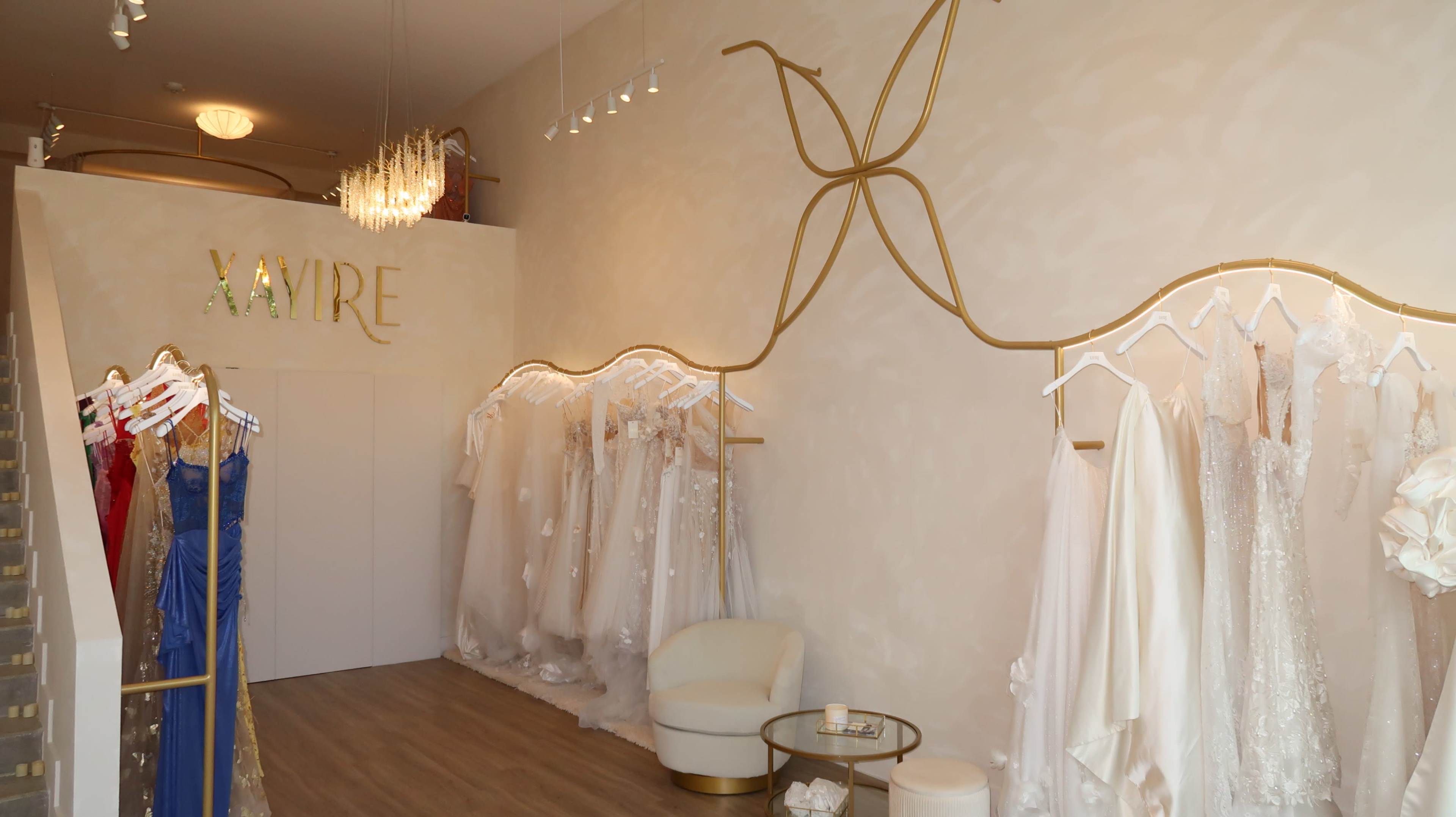 The image shows a bridal shop featuring various wedding dresses displayed on racks against a softly lit wall with a decorative gold design and a seating area.