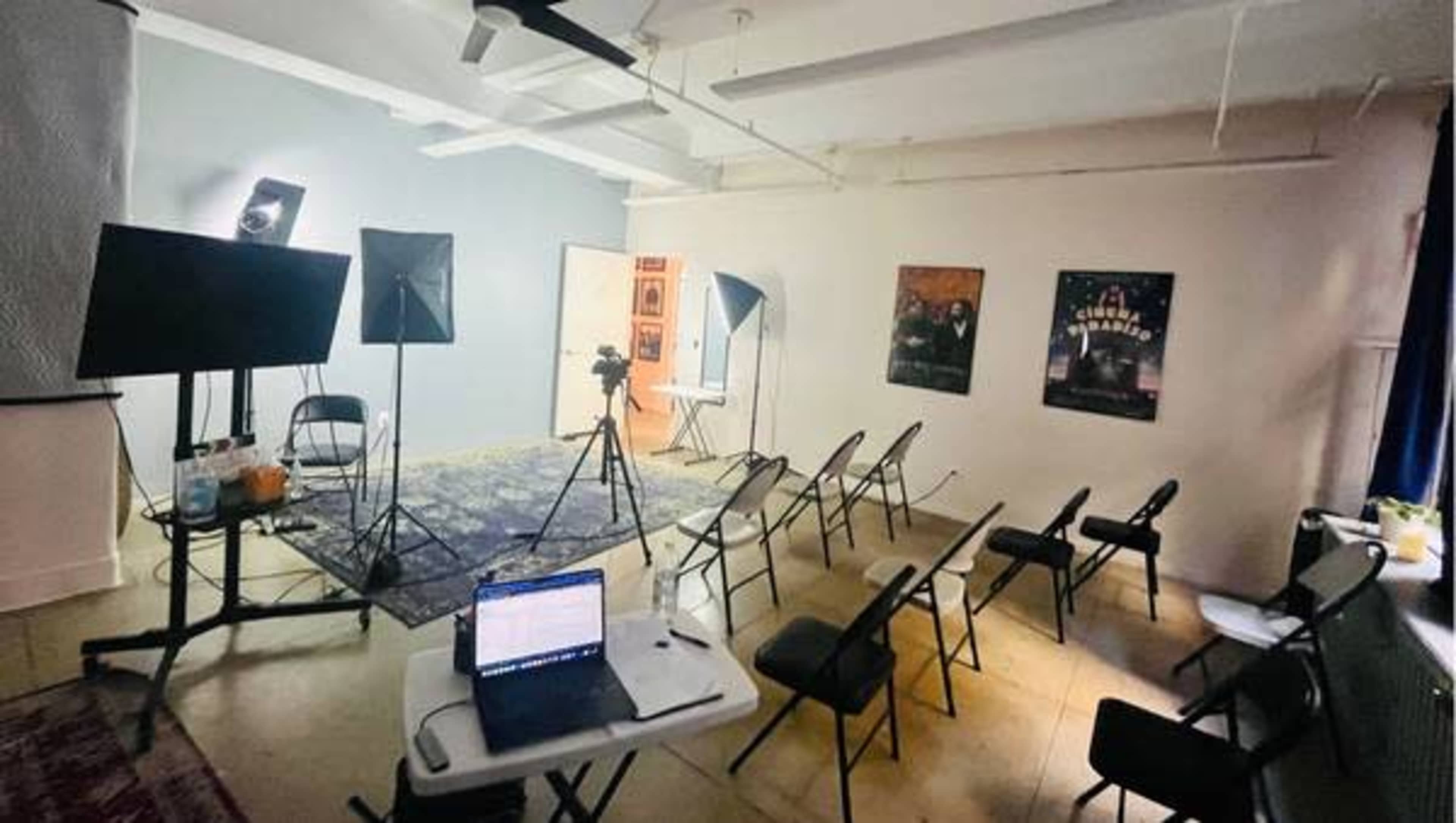 The image shows a spacious room set up for a presentation, featuring several chairs arranged in rows, a laptop on a table, and lighting equipment positioned for filming.