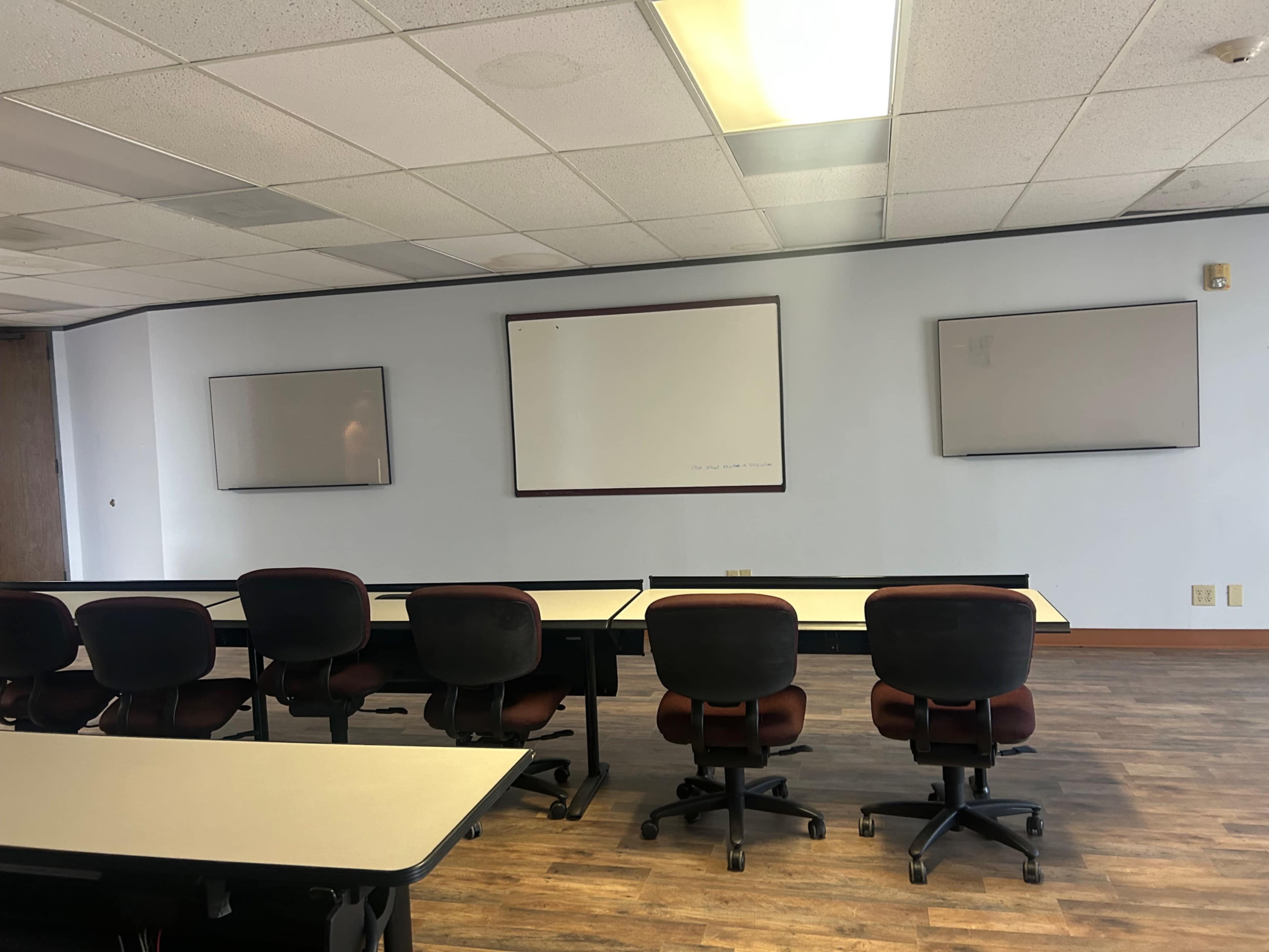 The image shows a conference room with multiple tables and chairs arranged in front of three wall-mounted whiteboards.
