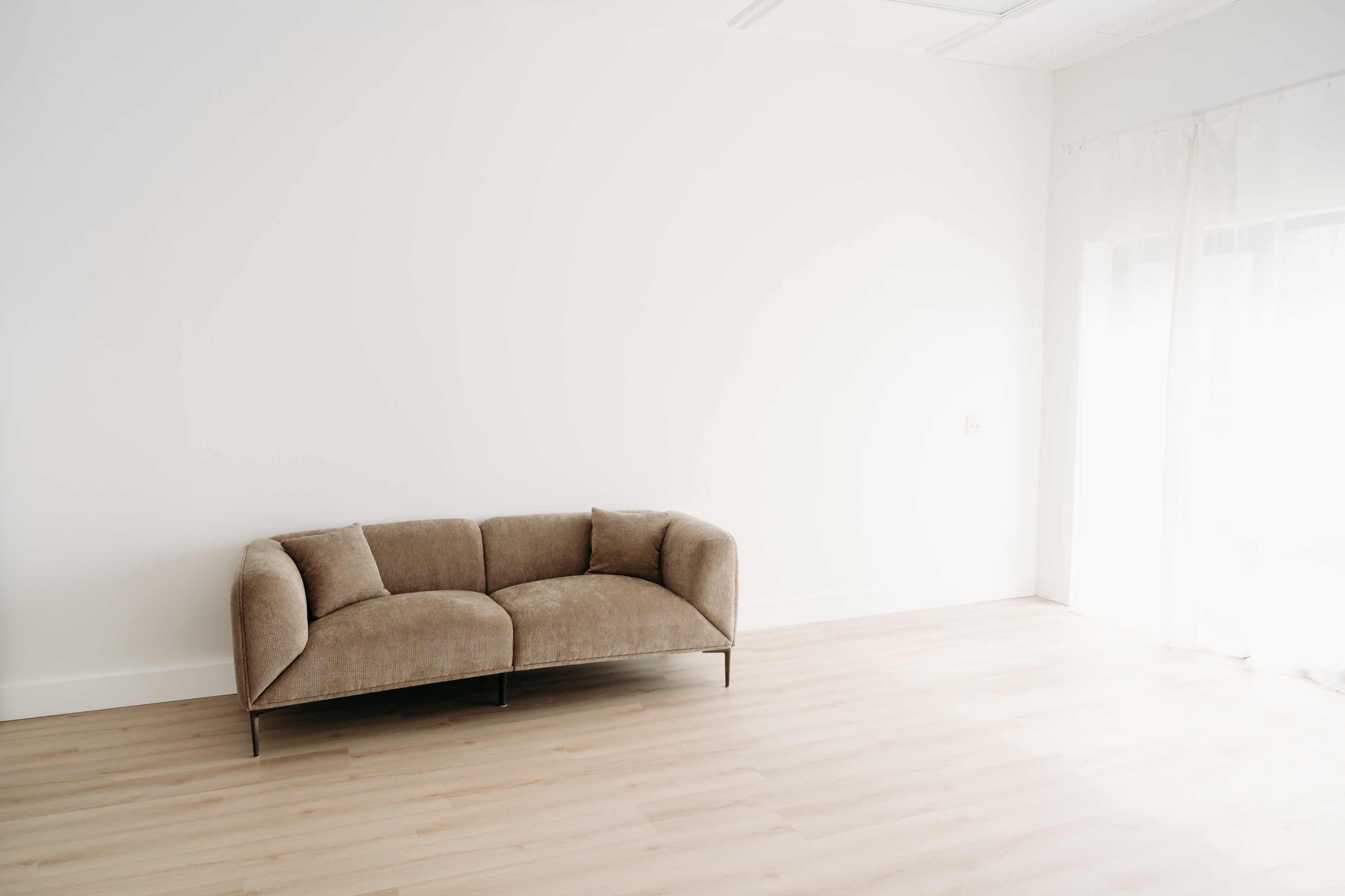 A beige tufted sofa sits against a plain white wall in a minimally furnished room with natural light streaming through a sheer curtain.