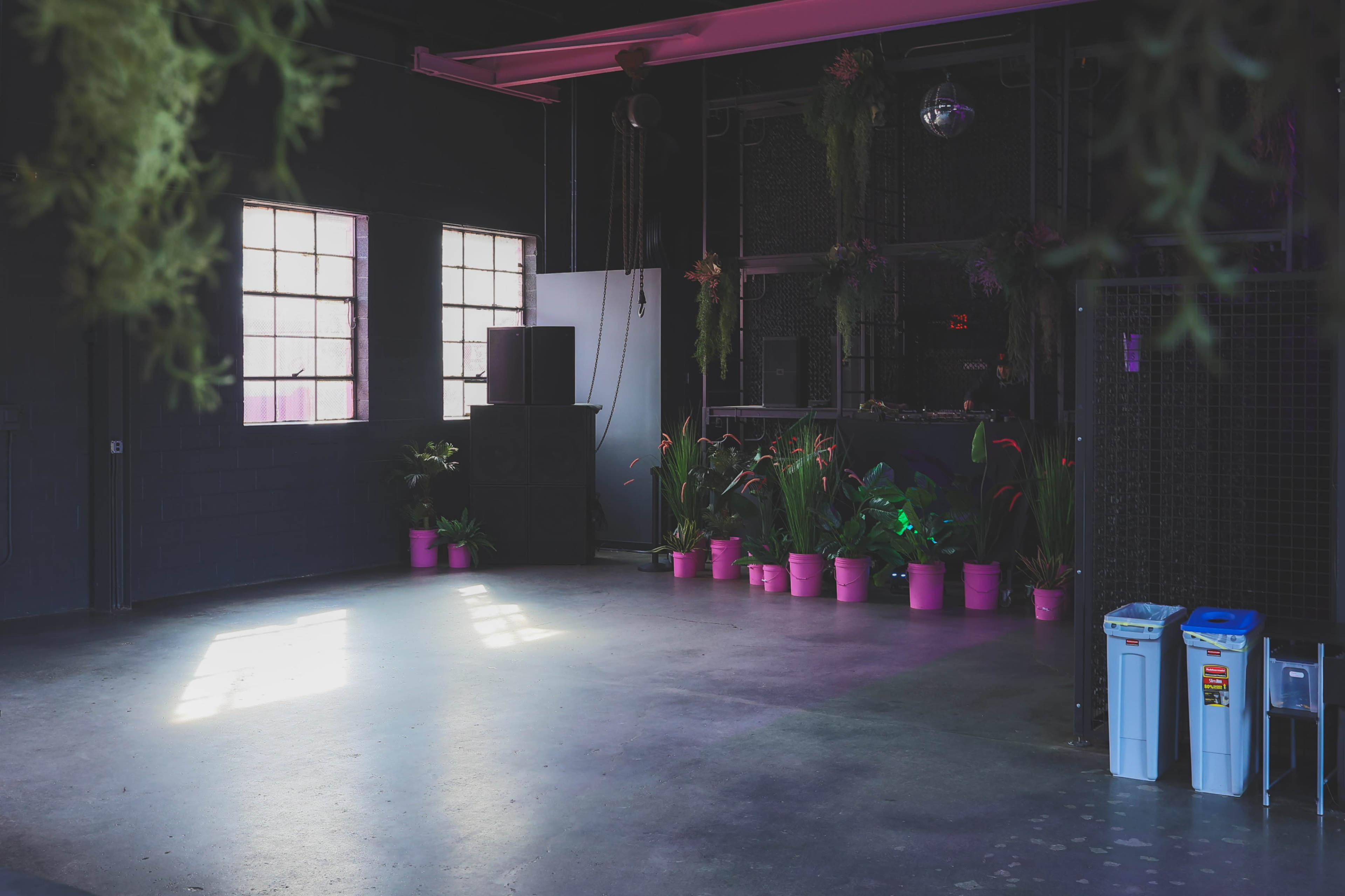 An empty indoor space with large windows, pink plant pots along the walls, and a sound system in the corner.
