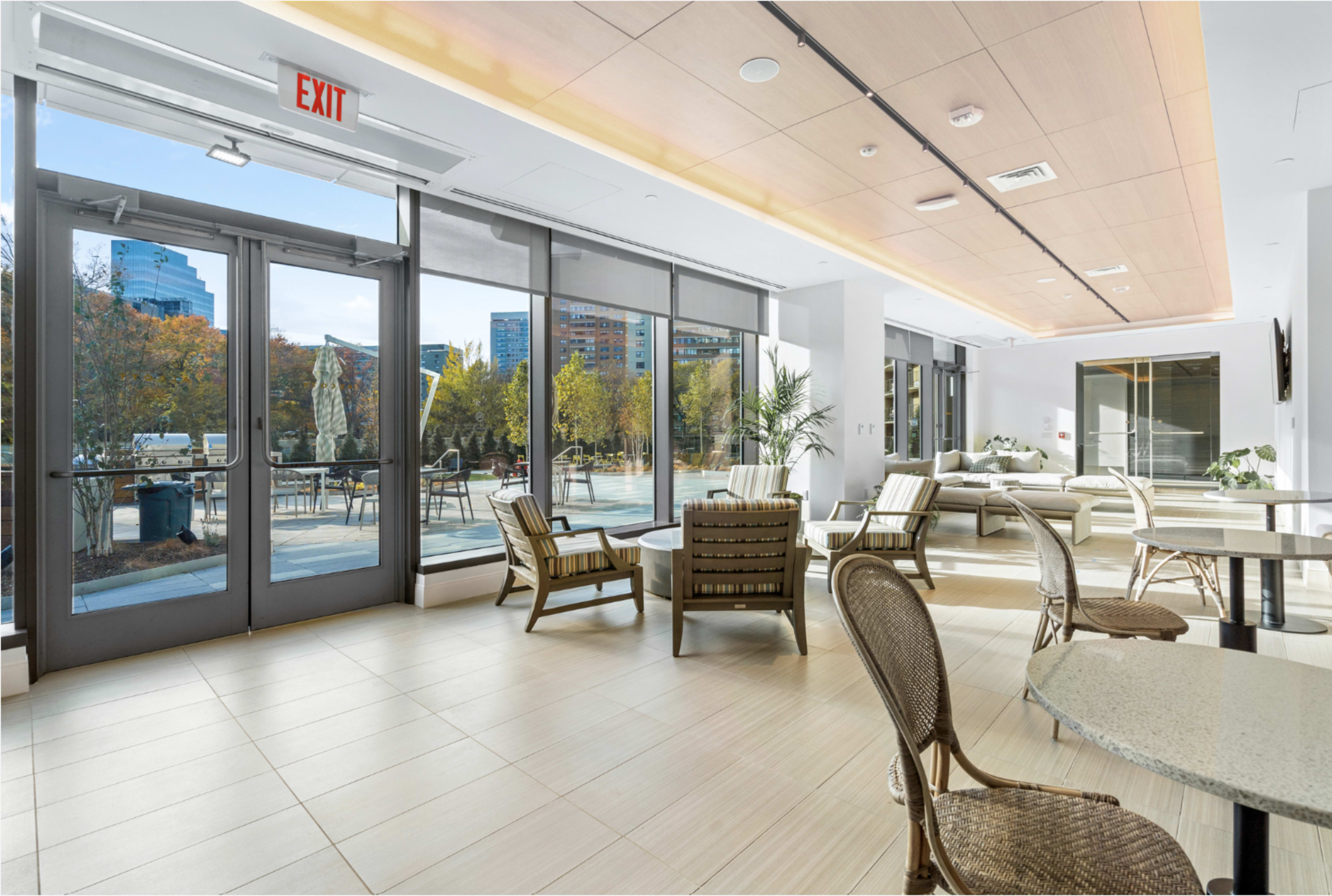 The image shows a modern, well-lit indoor space with floor-to-ceiling windows, showcasing a view of an outdoor area with trees and buildings in the background.