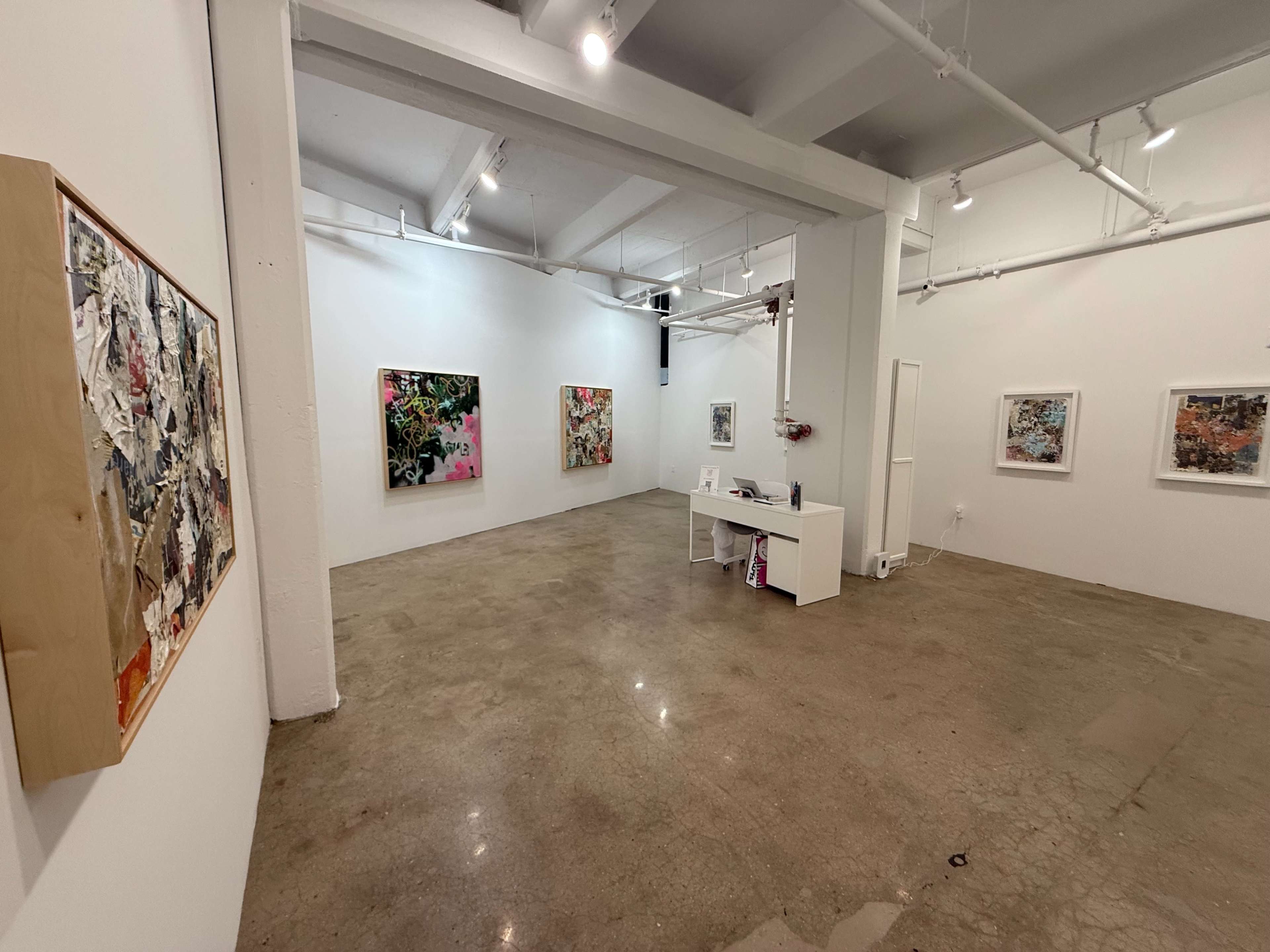 The image shows an art gallery interior with several colorful abstract paintings displayed on the walls and a white desk positioned in the foreground.