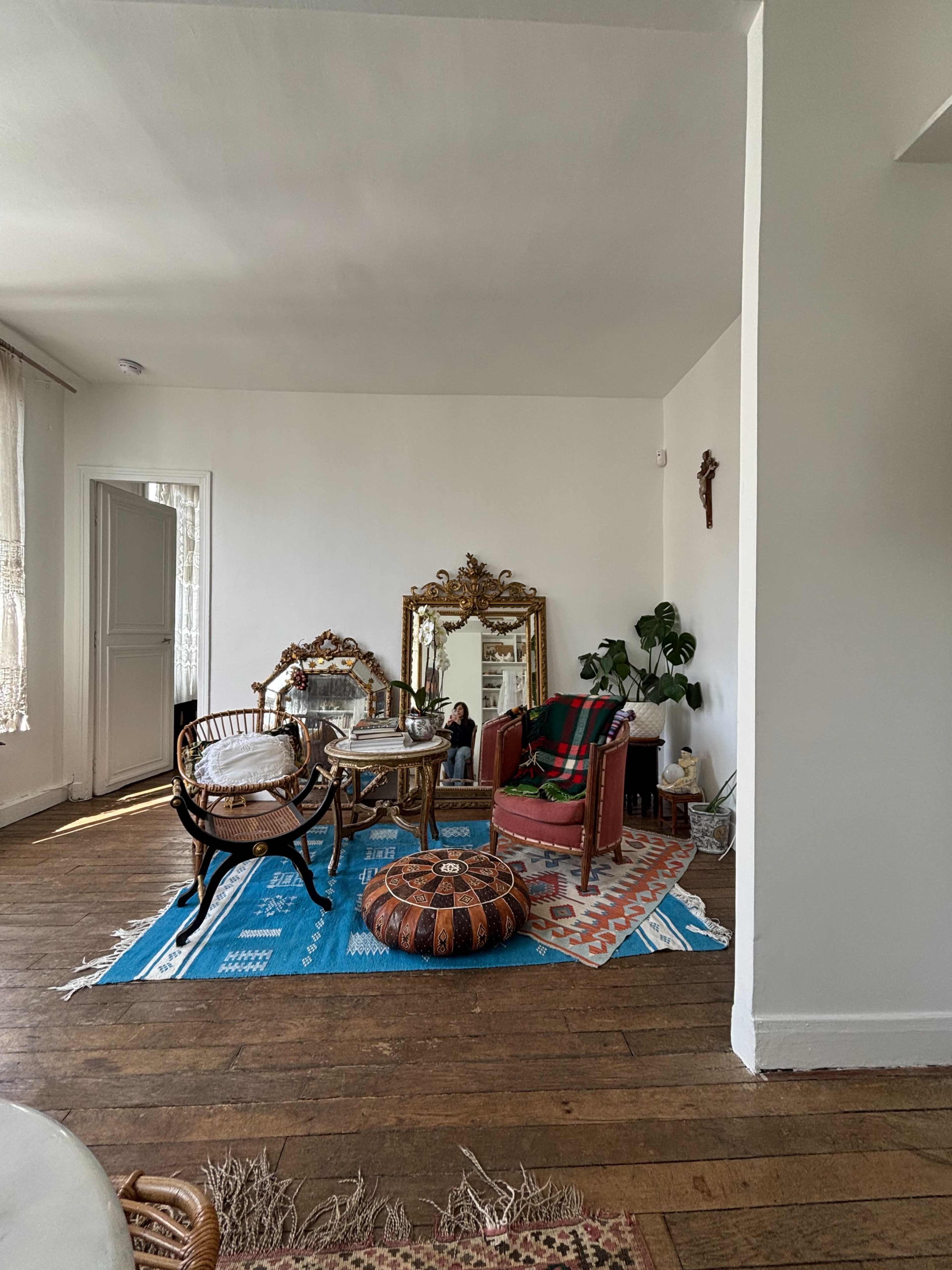 The image depicts a bright living room featuring a mix of vintage furniture, a large mirror, and colorful rugs on wooden flooring.
